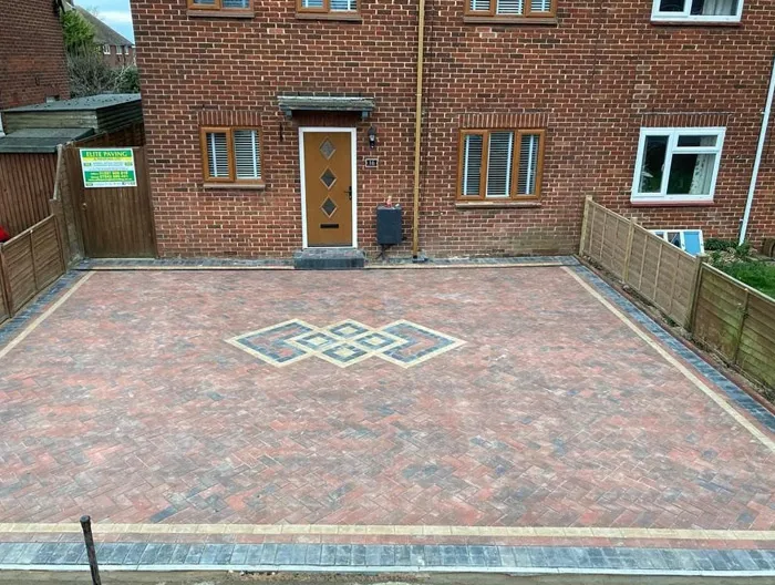 A pristine new block paved driveway featuring red and grey blocks with an intricate geometric pattern in the centre, bordered by yellow and dark grey blocks, in front of a red brick house with a brown front door.