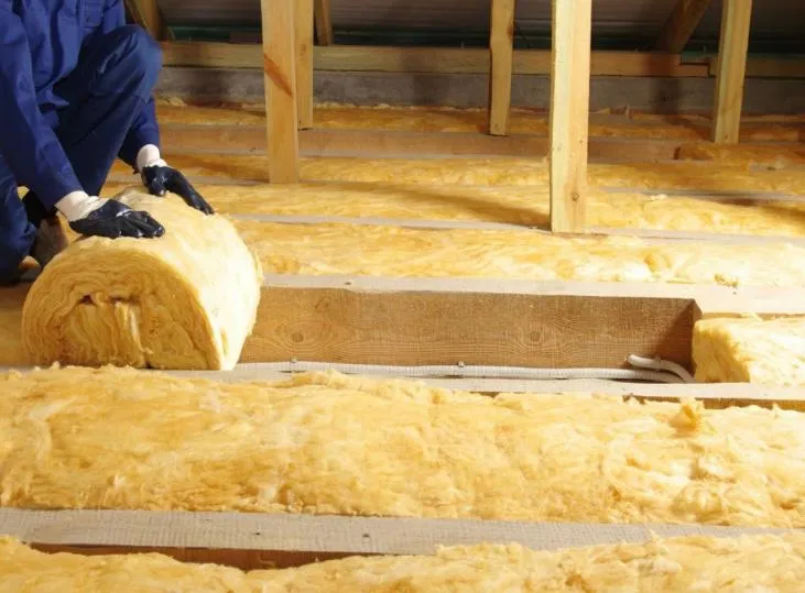Worker in blue overalls and gloves unrolling and installing yellow mineral wool insulation in a loft.