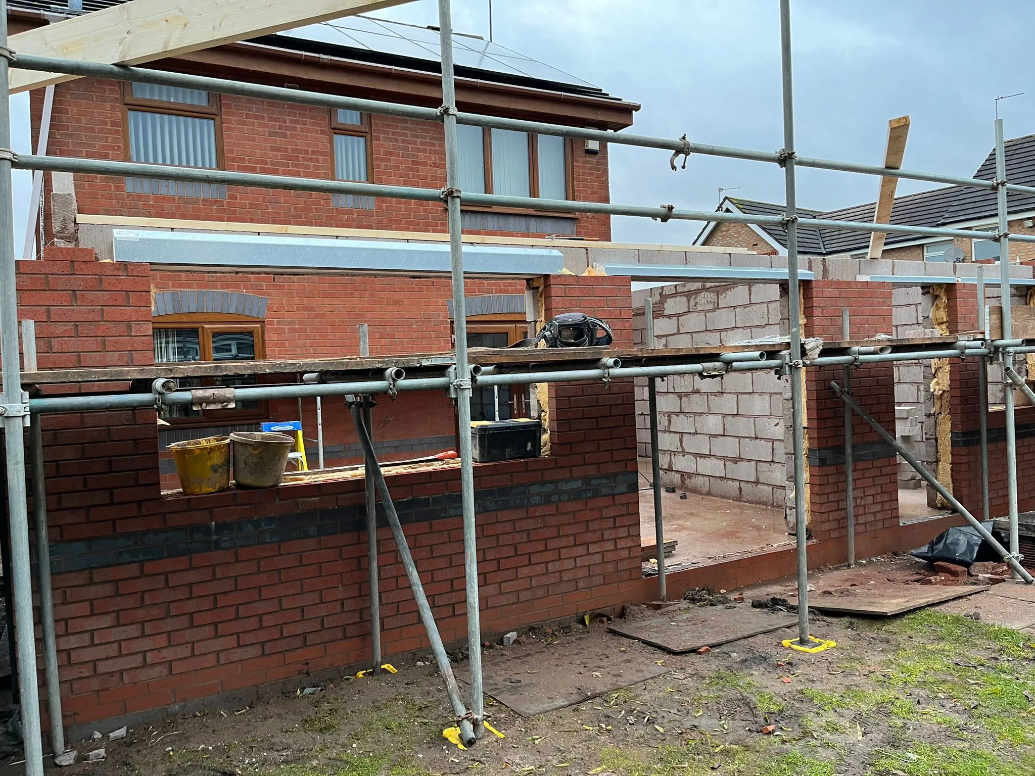 An active construction site for a home extension, featuring scaffolding, red brickwork, concrete blocks, and tools.