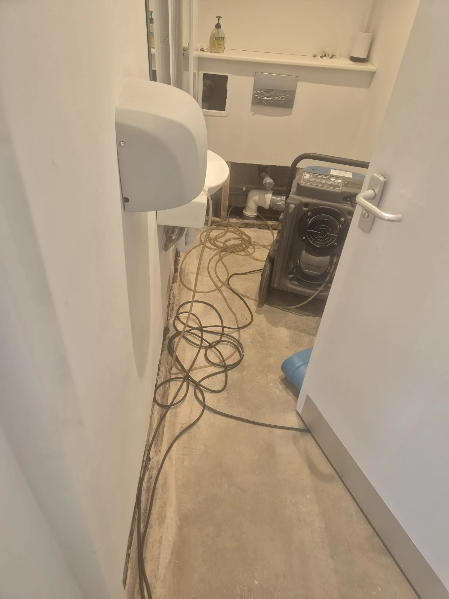 Commercial washroom undergoing refurbishment with visible pipes, wiring, a hand dryer, and a dehumidifier on the floor.