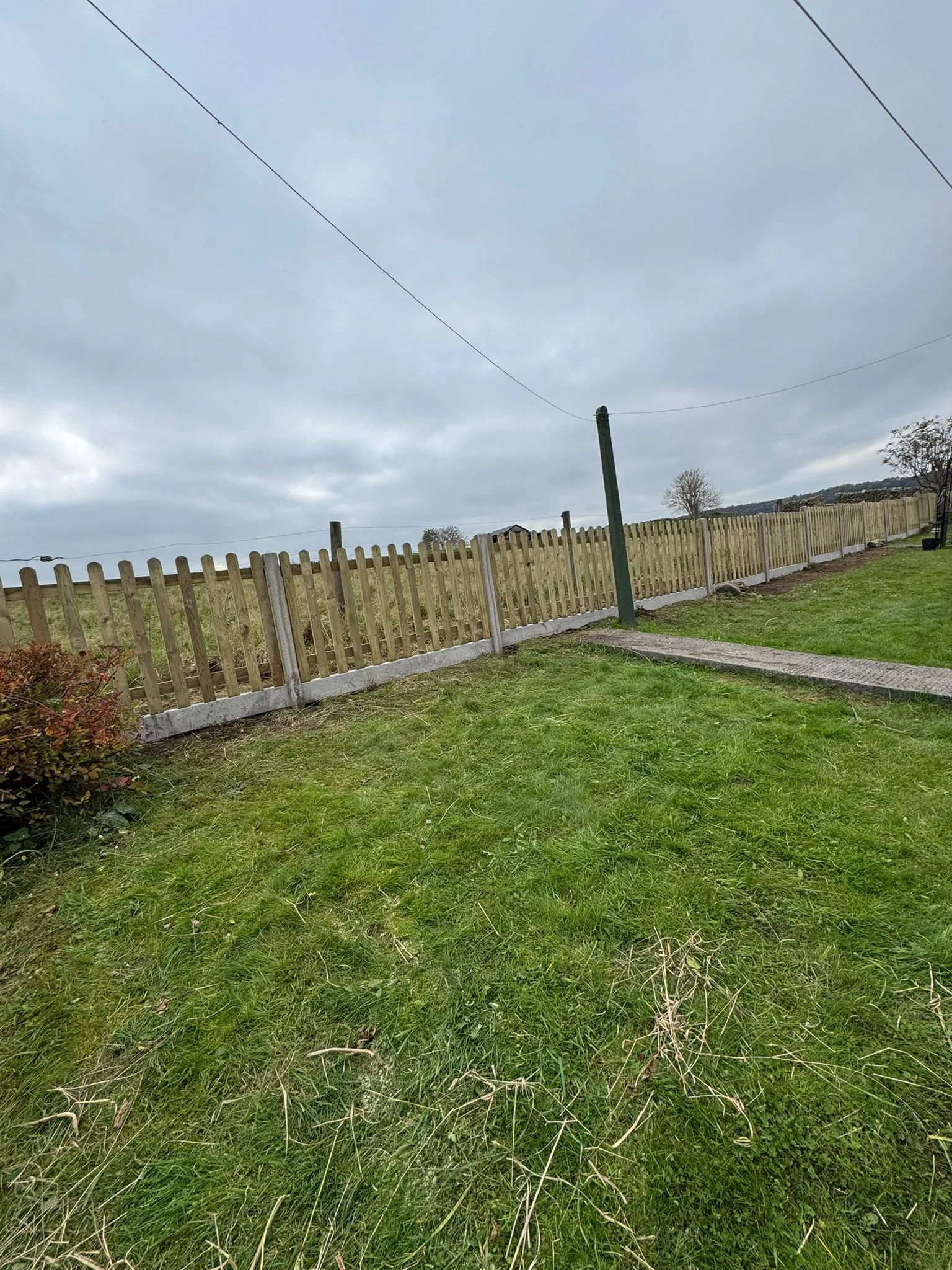 A long wooden picket fence with concrete posts and gravel boards separating a lawn from a field.