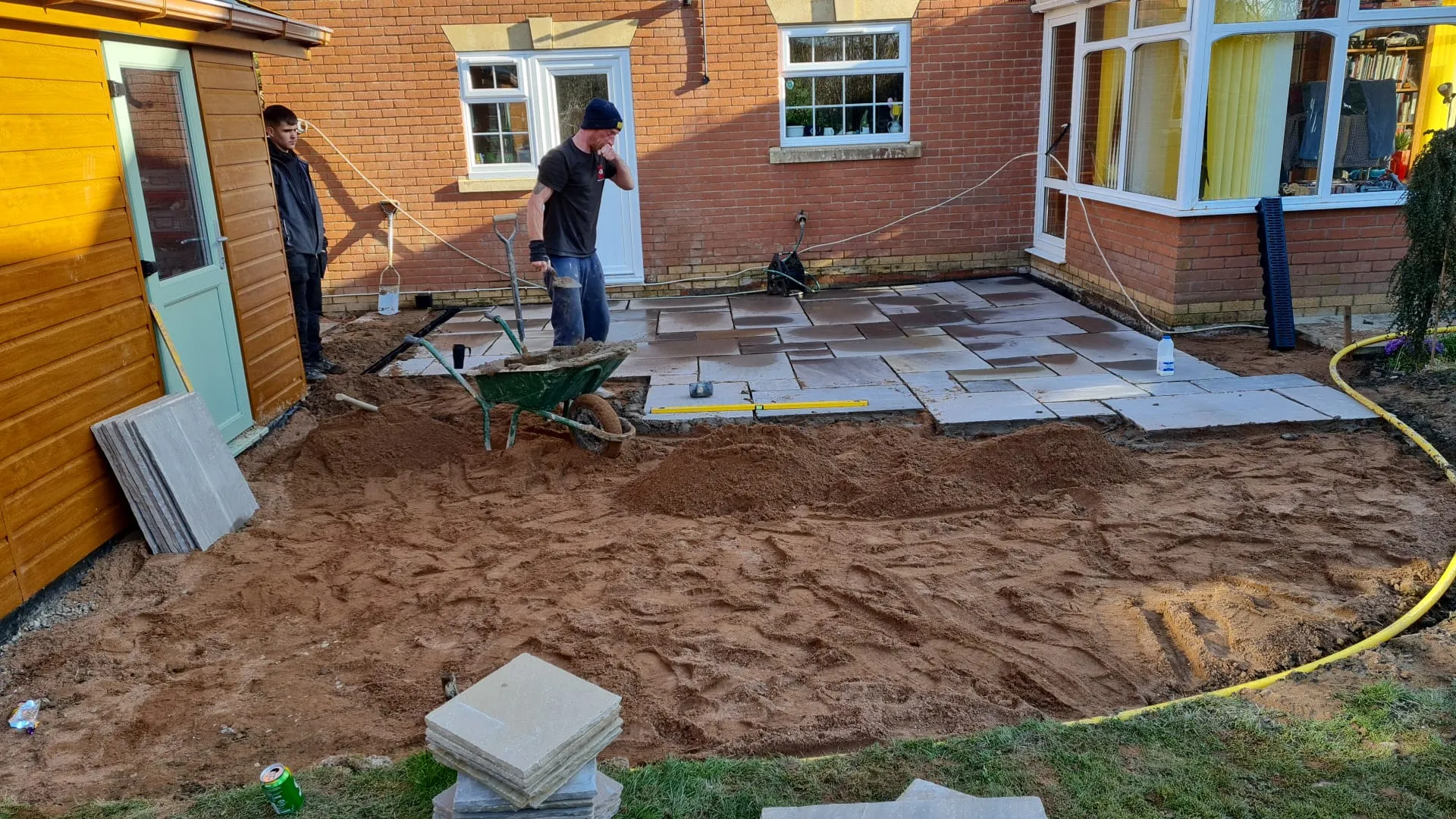 A high-quality image of two workers laying a new patio with large natural stone slabs in a residential garden, showing the progress of the installation with sand base and materials.