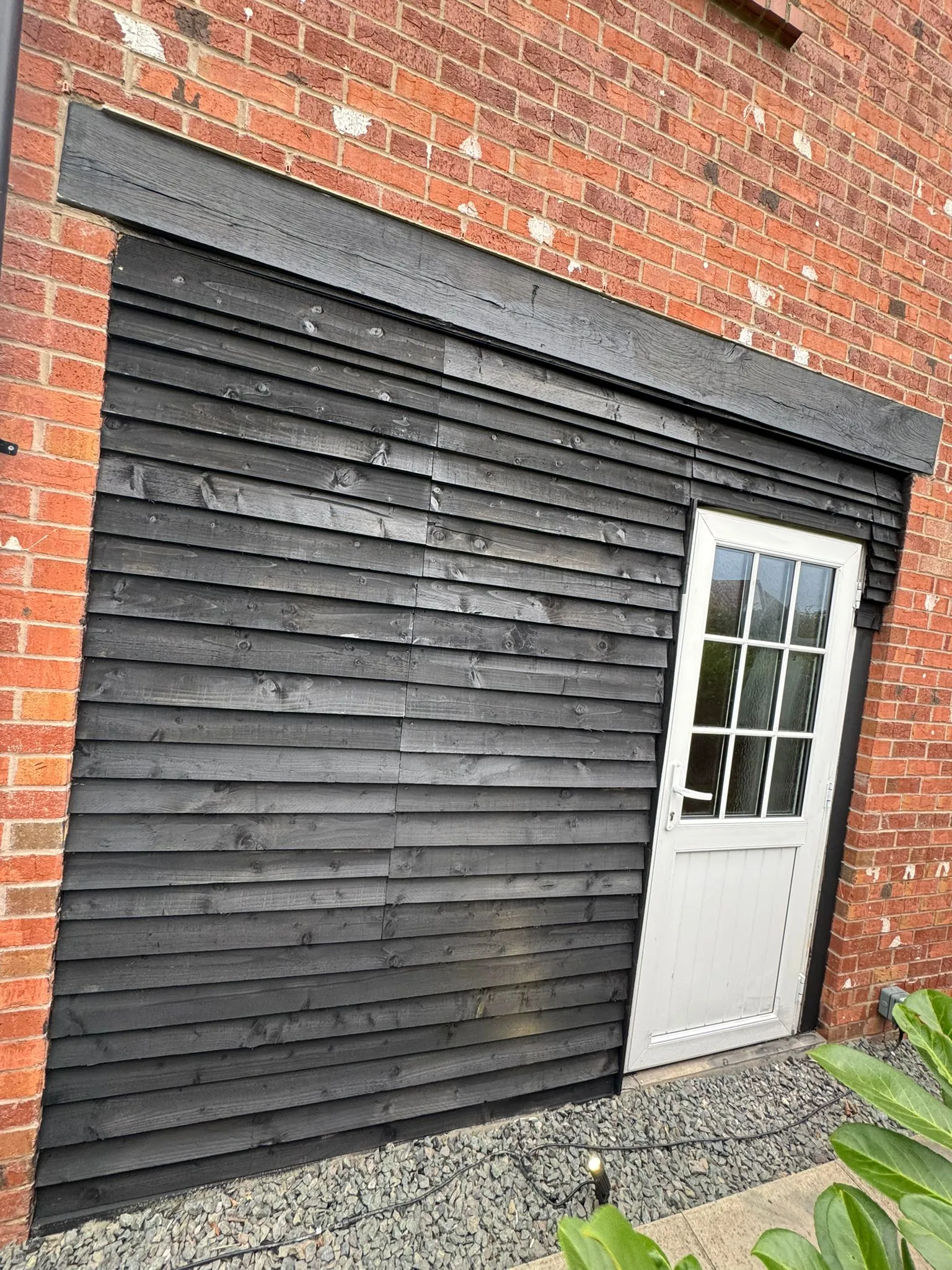 Close-up of a house exterior with dark wooden cladding, red brickwork, and a white door, showcasing exterior property renovation or maintenance.