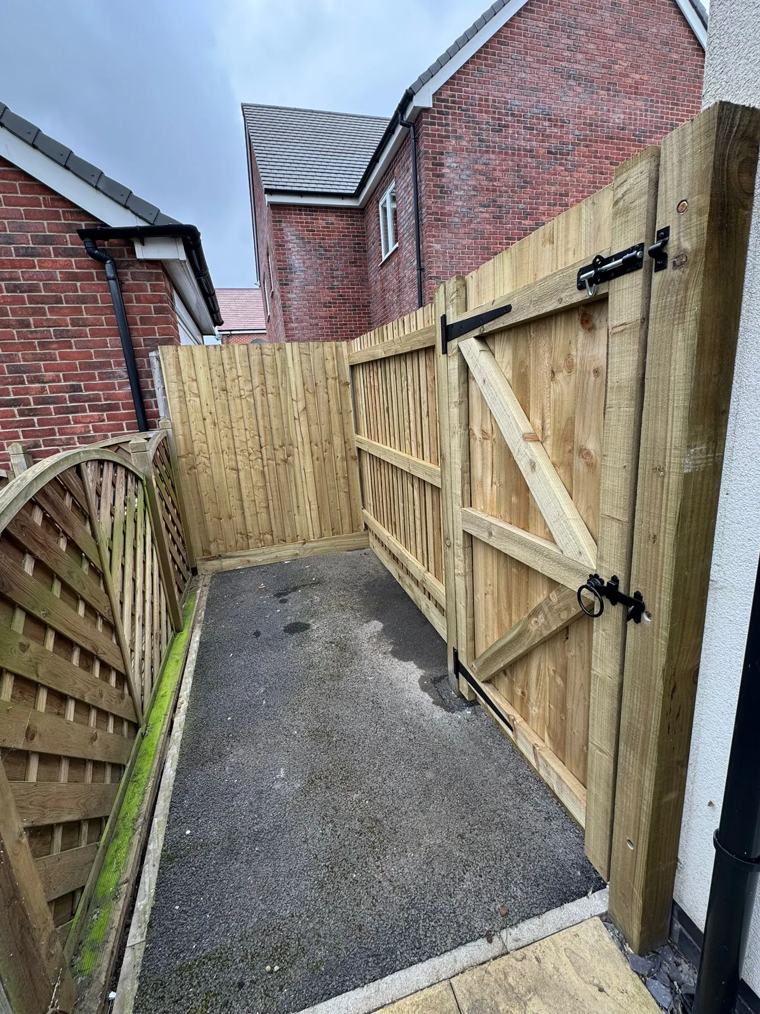 A close-up of a recently installed wooden garden fence and gate with black hardware, next to an asphalt path and brick houses.