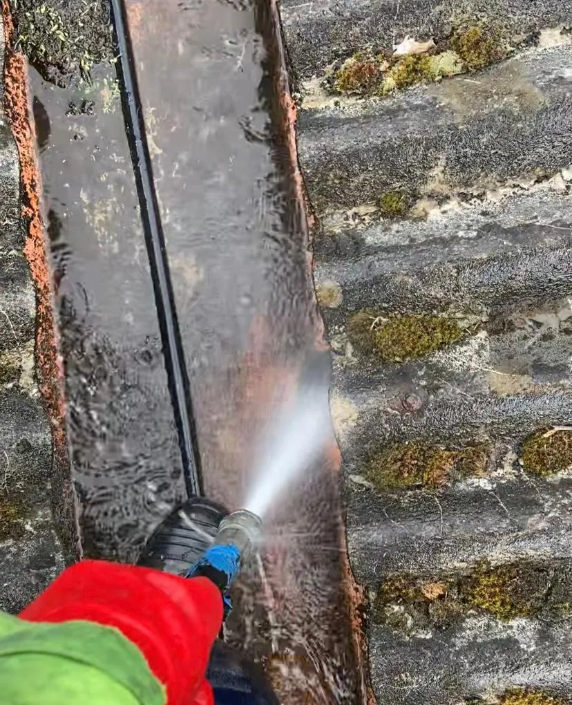 High-pressure water jet being used to clean a gully filled with dirty water and sediment, with moss on the surrounding surfaces.