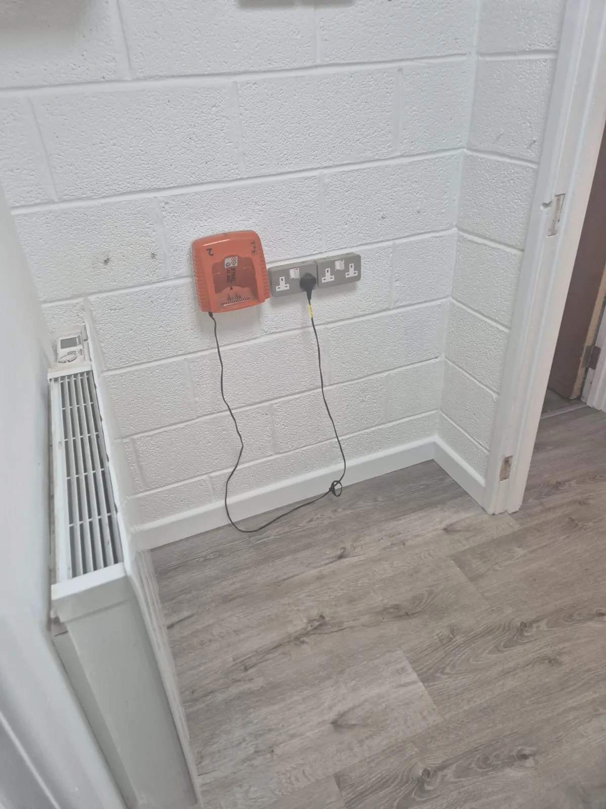 Interior of a refurbished space with white painted blockwork walls, new grey wood-effect laminate flooring, white skirting boards, a white radiator, an orange fire alarm call point, and two grey double electrical sockets.