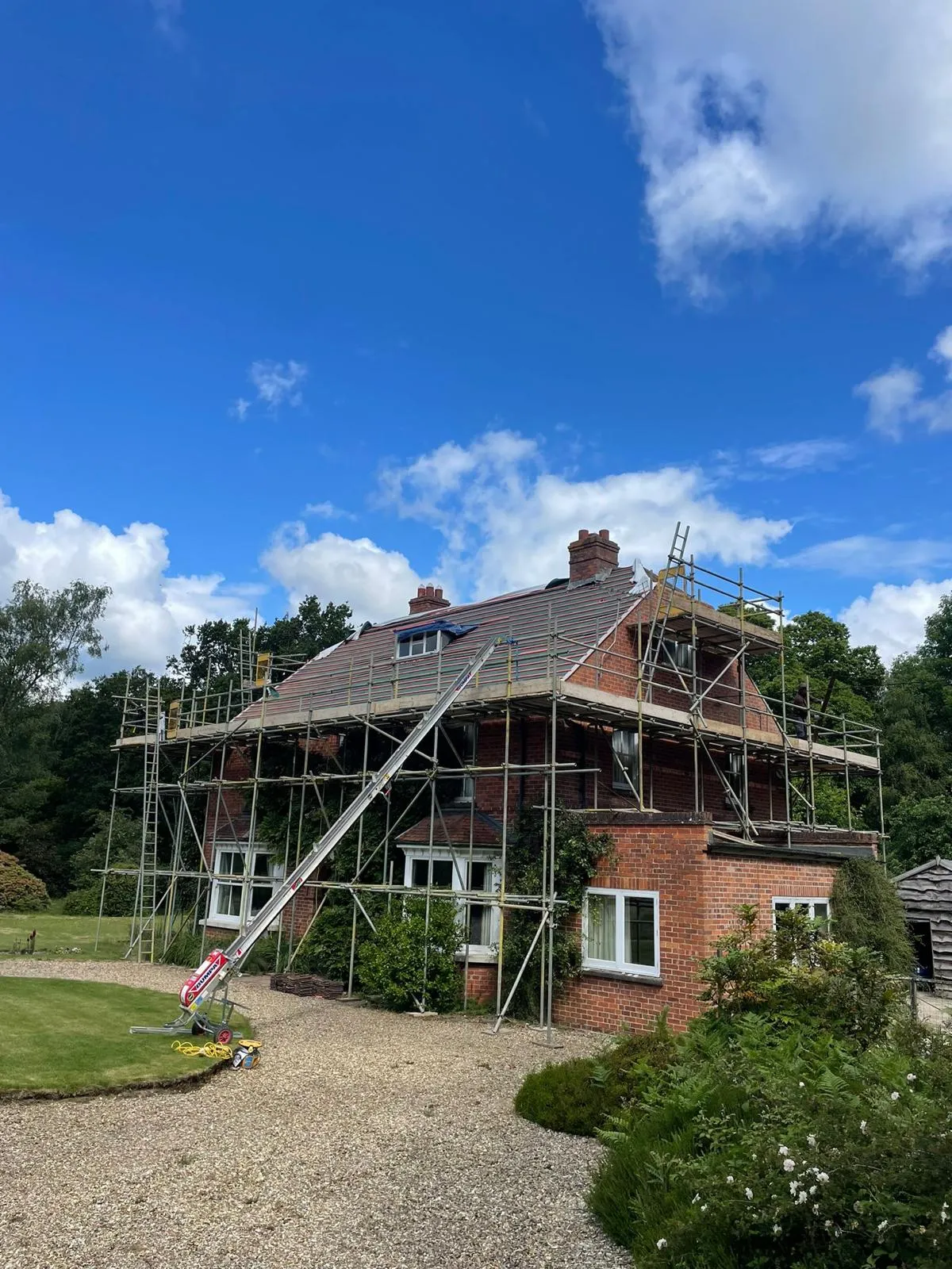 A red brick house covered in scaffolding with a new roof being installed, showing a tile hoist and the ongoing work.