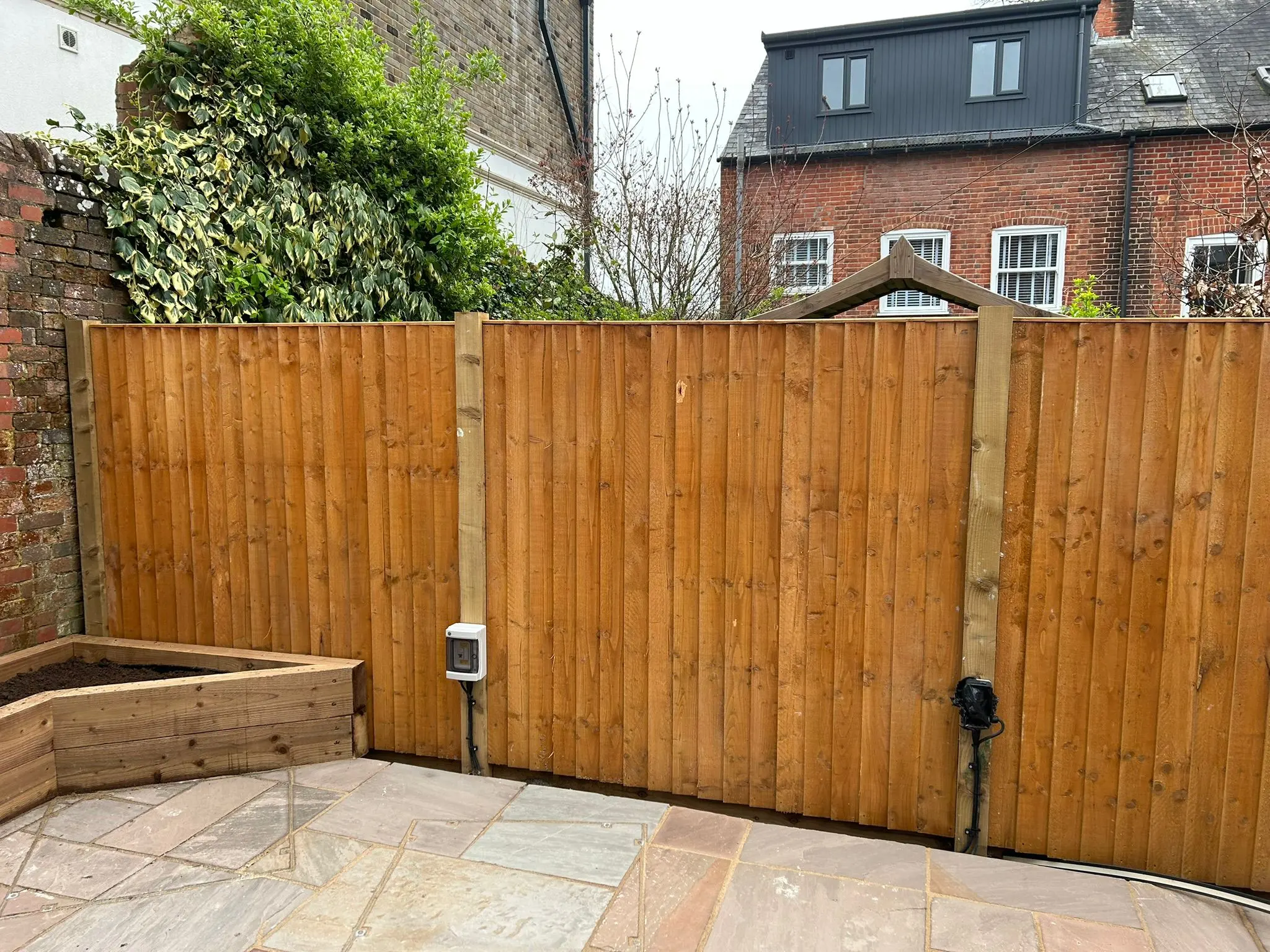 A close-up of a new, sturdy wooden garden fence installed in a residential backyard, featuring vertical wooden planks and robust wooden posts. The fence is situated adjacent to a light-coloured stone patio and a raised wooden planter. Electrical outlets are visible on the fence posts.