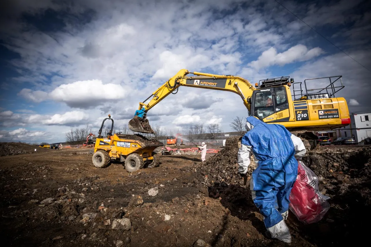 Commercial asbestos removal on a large construction site with an excavator, a dumper, and workers in blue and white hazmat suits managing debris and hazardous waste.