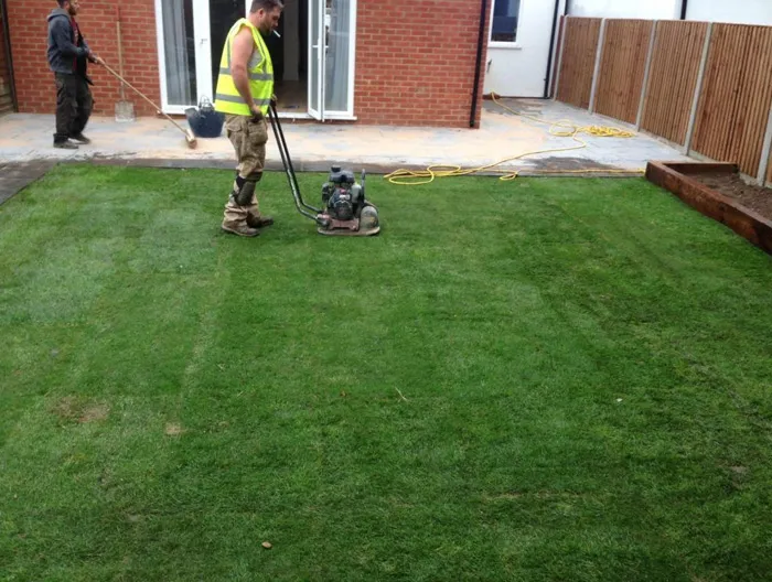 Landscapers laying and compacting a new turf lawn with a wacker plate next to a modern patio and wooden fence.
