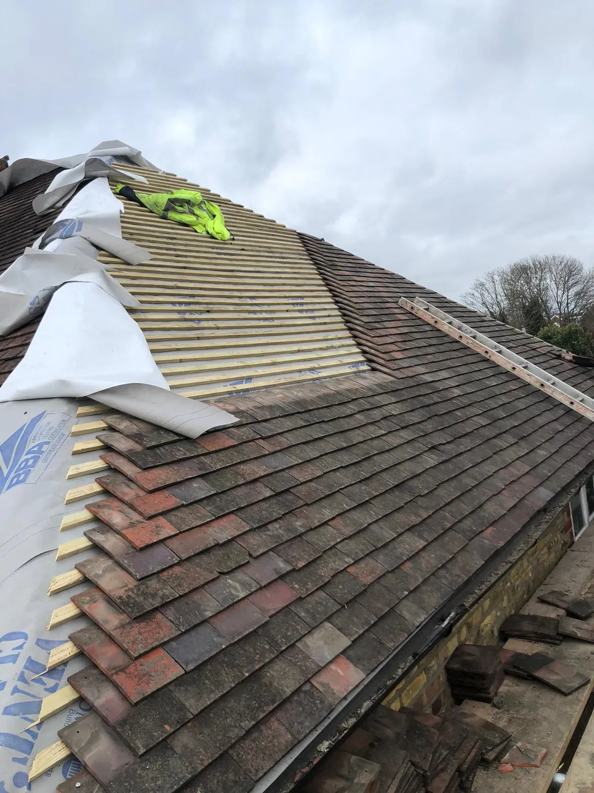 Re-roofing in progress on a pitched roof, displaying old roof tiles alongside new roofing felt (underlay) and timber battens, with a ladder leaning against the roof.