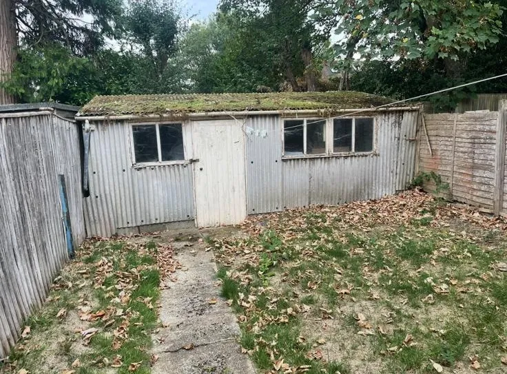 An old garden outbuilding with moss-covered corrugated asbestos cement roofing and walls in a residential garden.