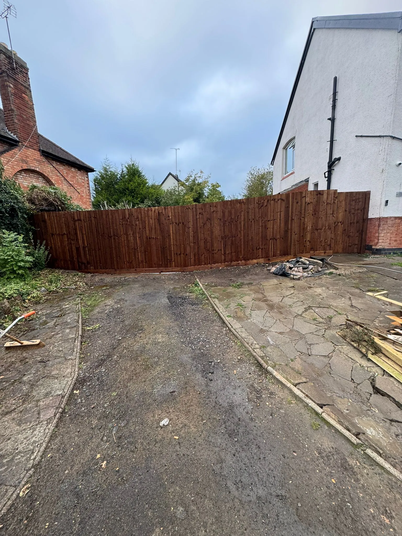 A newly installed dark wooden closeboard fence in a garden that is being renovated, with old paving and an unpaved section in the foreground.