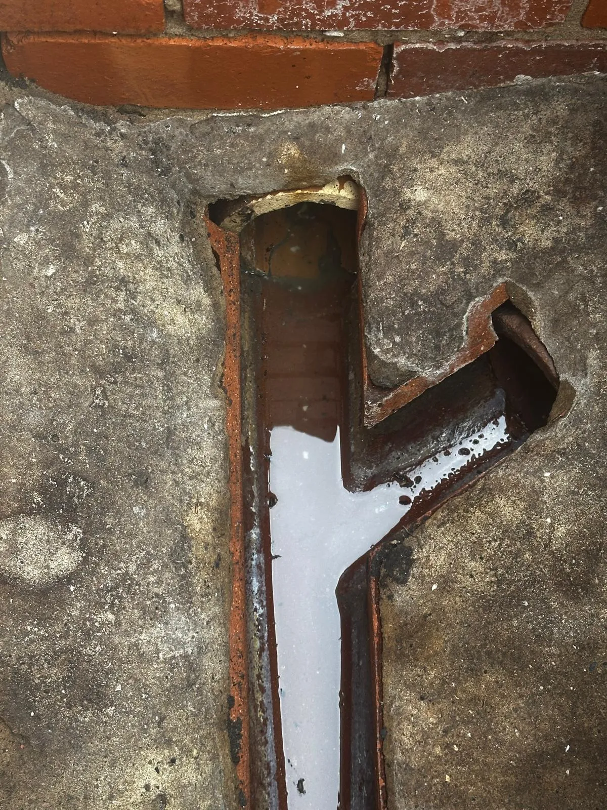 Close-up of a damaged and blocked outdoor gully drain overflowing with standing water near a brick wall.