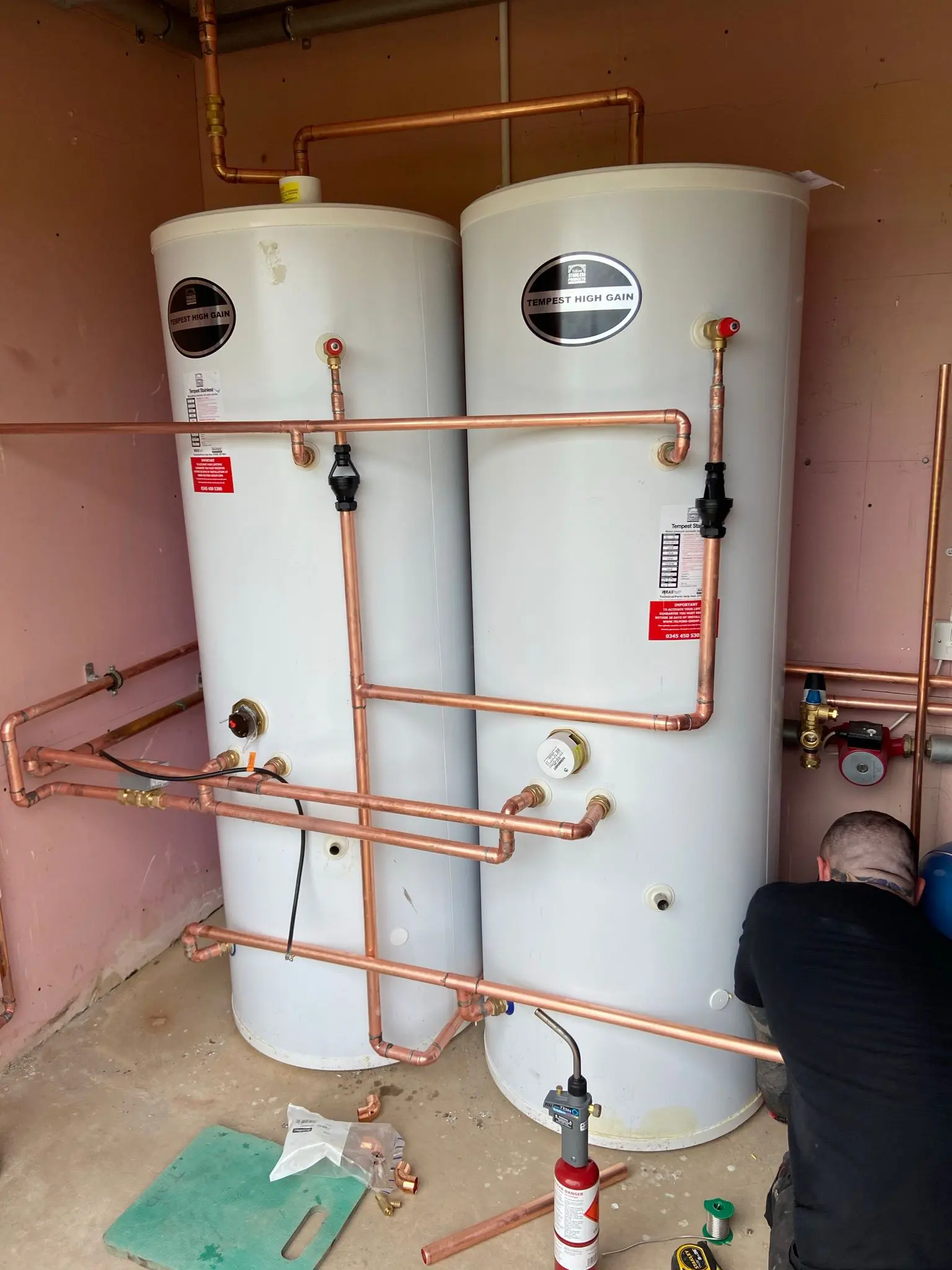Fact Services engineer installing two large white unvented hot water cylinders with complex copper pipework in a commercial or large residential property.