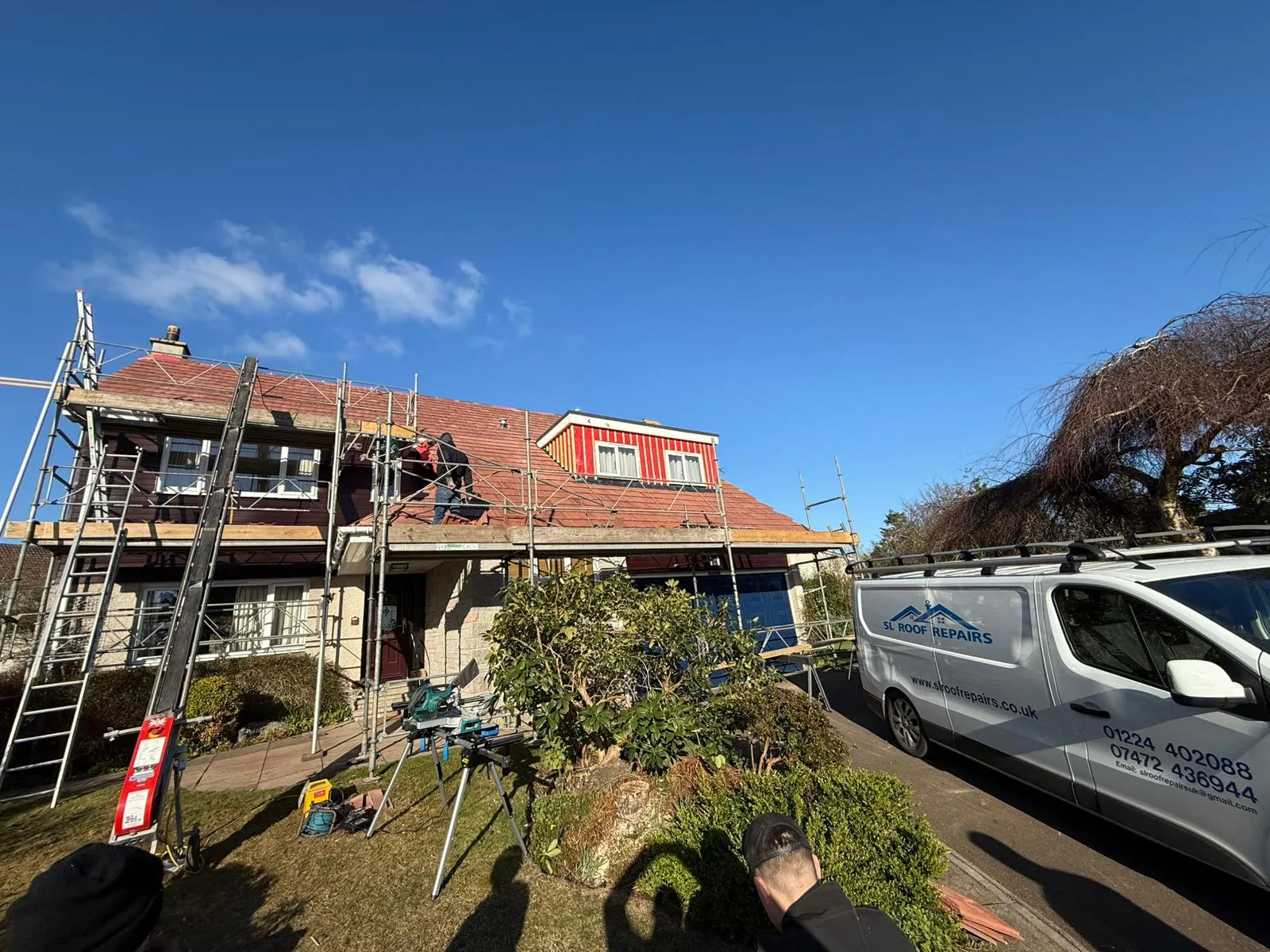 Roofers on scaffolding working on a house's red-tiled roof and dormer window, with a professional work van parked outside.