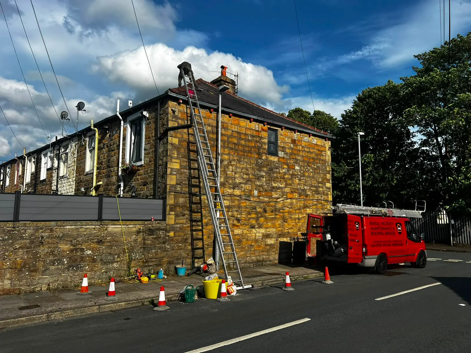 Roofer on ladder repairing stone house roof, with red company van and safety cones below.