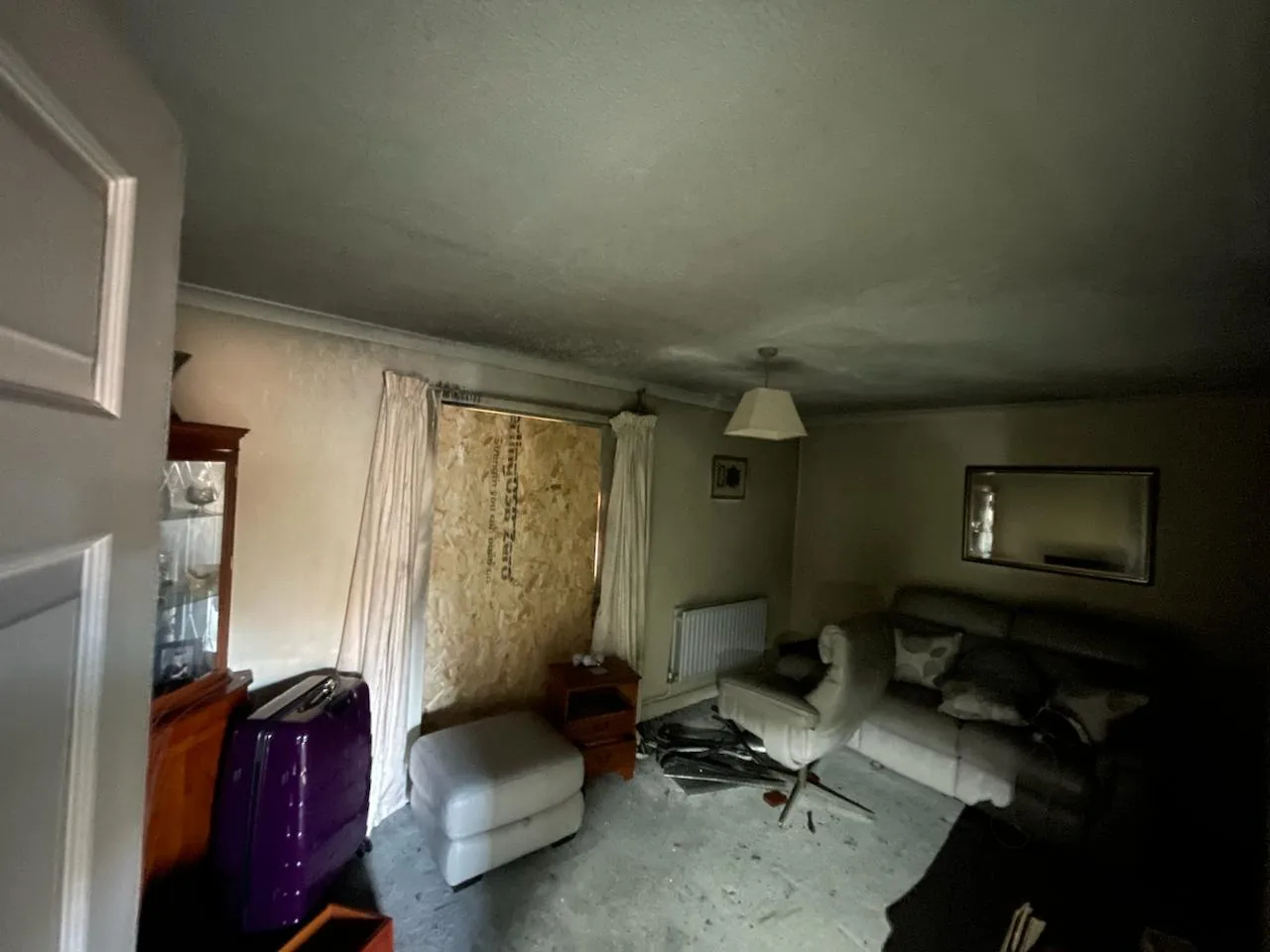 Interior of a damaged living room with a textured ceiling, boarded window, and furniture. The ceiling shows signs of discolouration or smoke damage.
