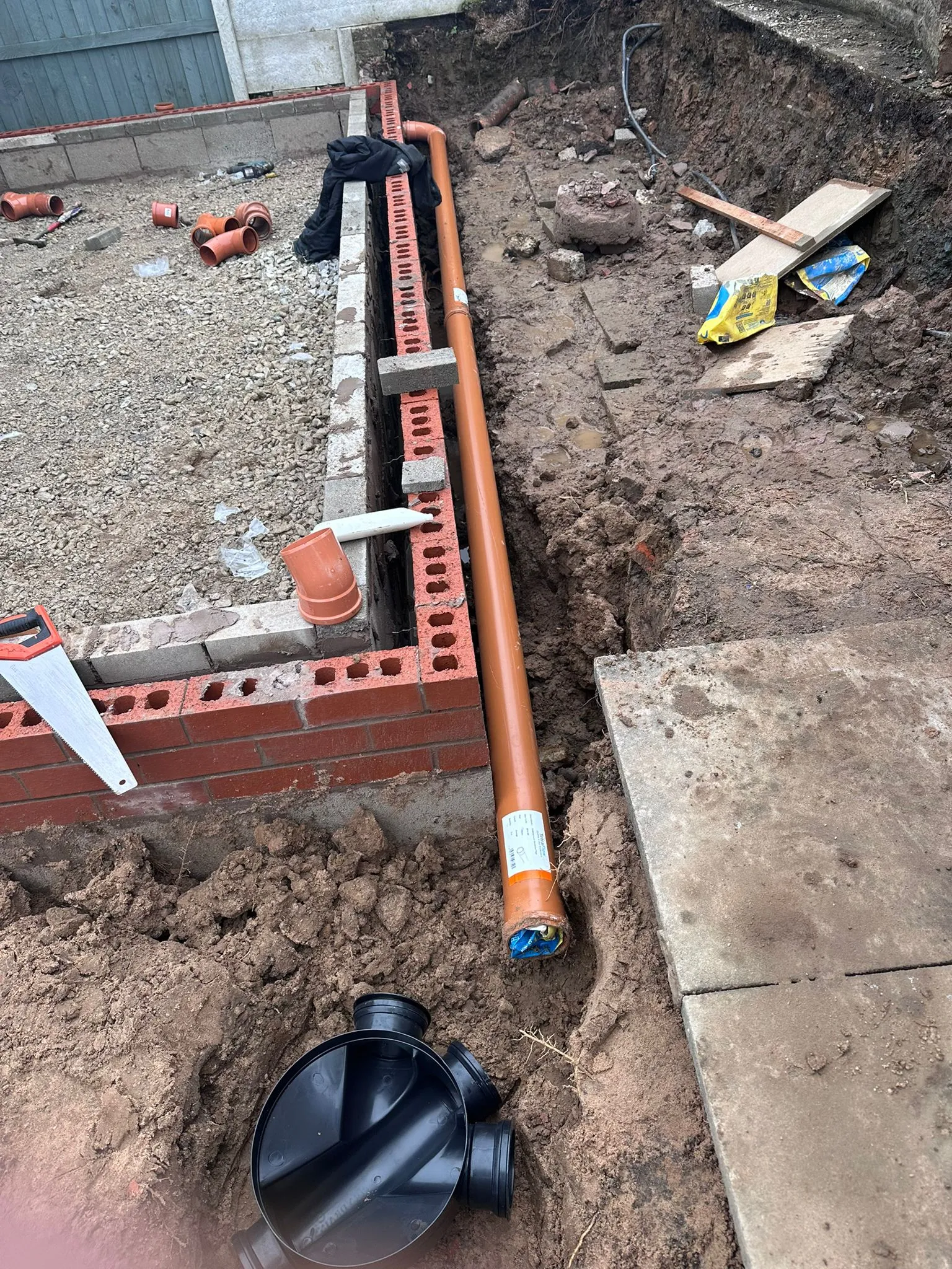 New orange drainage pipes and a black plastic inspection chamber being installed in an excavated trench alongside new brick footings.