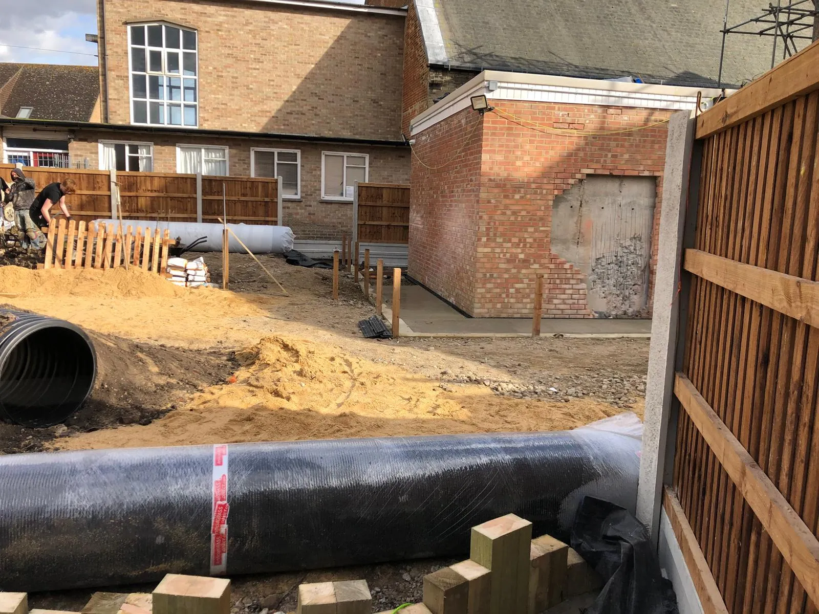 Construction site with groundworks, large black drainage pipes, and a partially built brick extension, demonstrating the early stages of a building project.