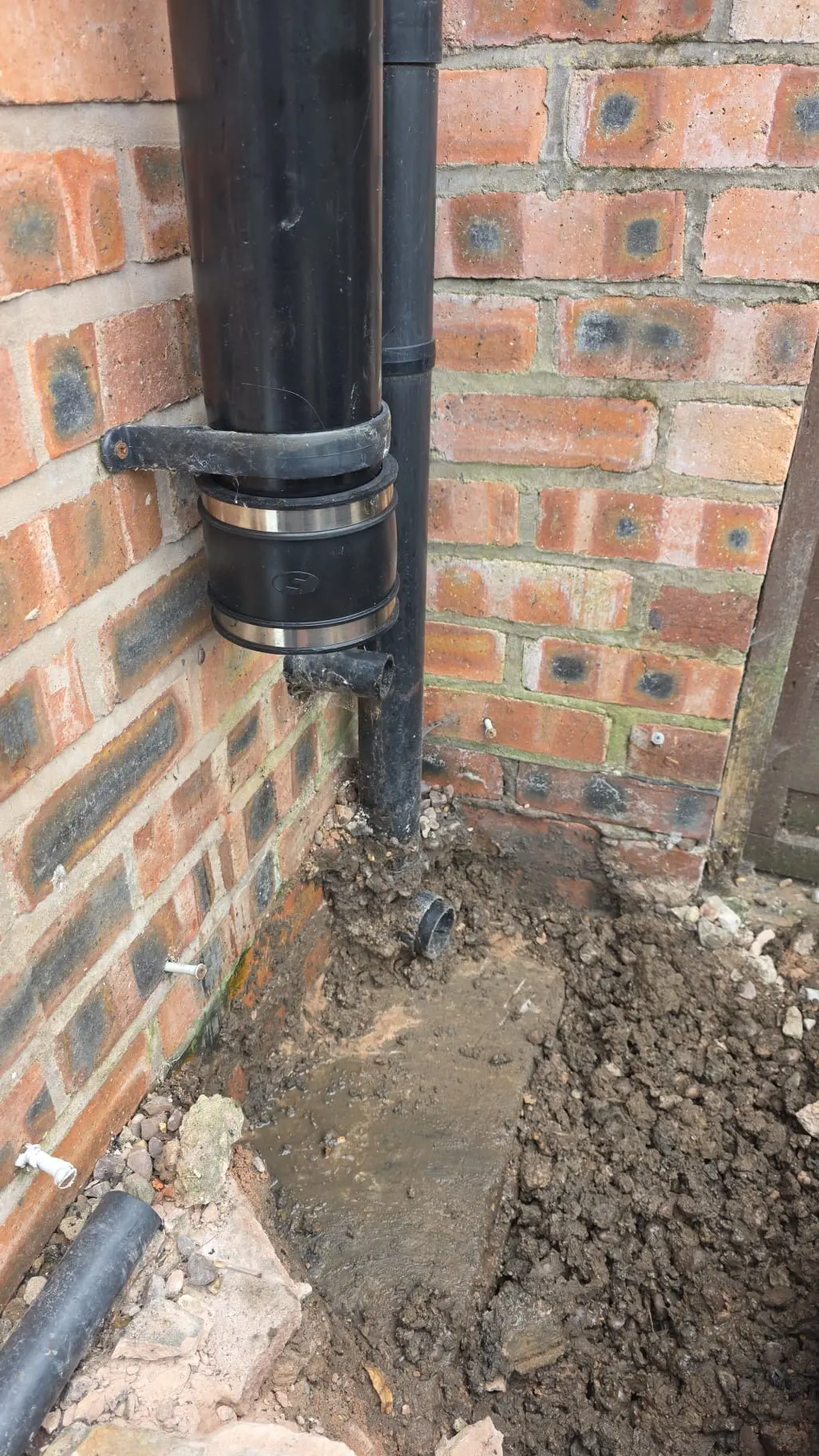 Excavated external drain pipe and muddy groundwork against a residential brick wall