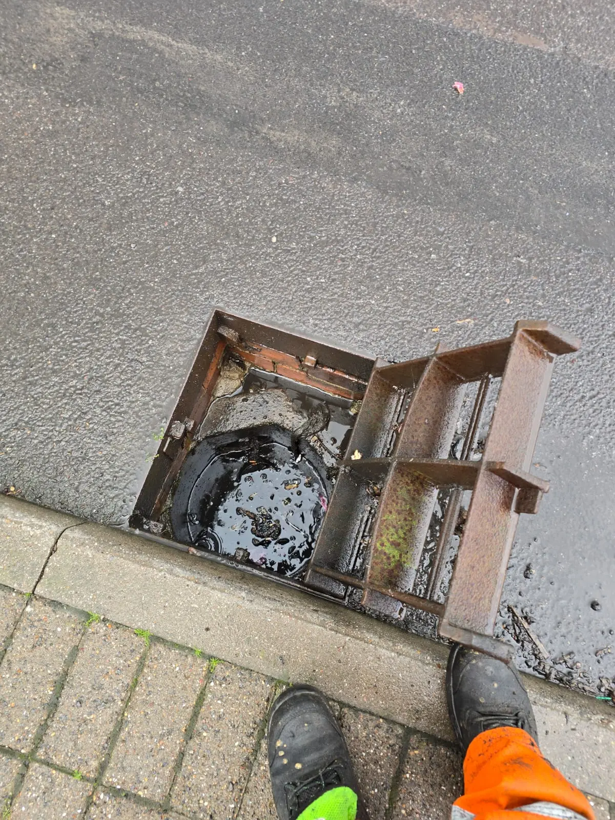 A drainage engineer inspecting an open, blocked street drain full of dirty standing water next to a paved curb.