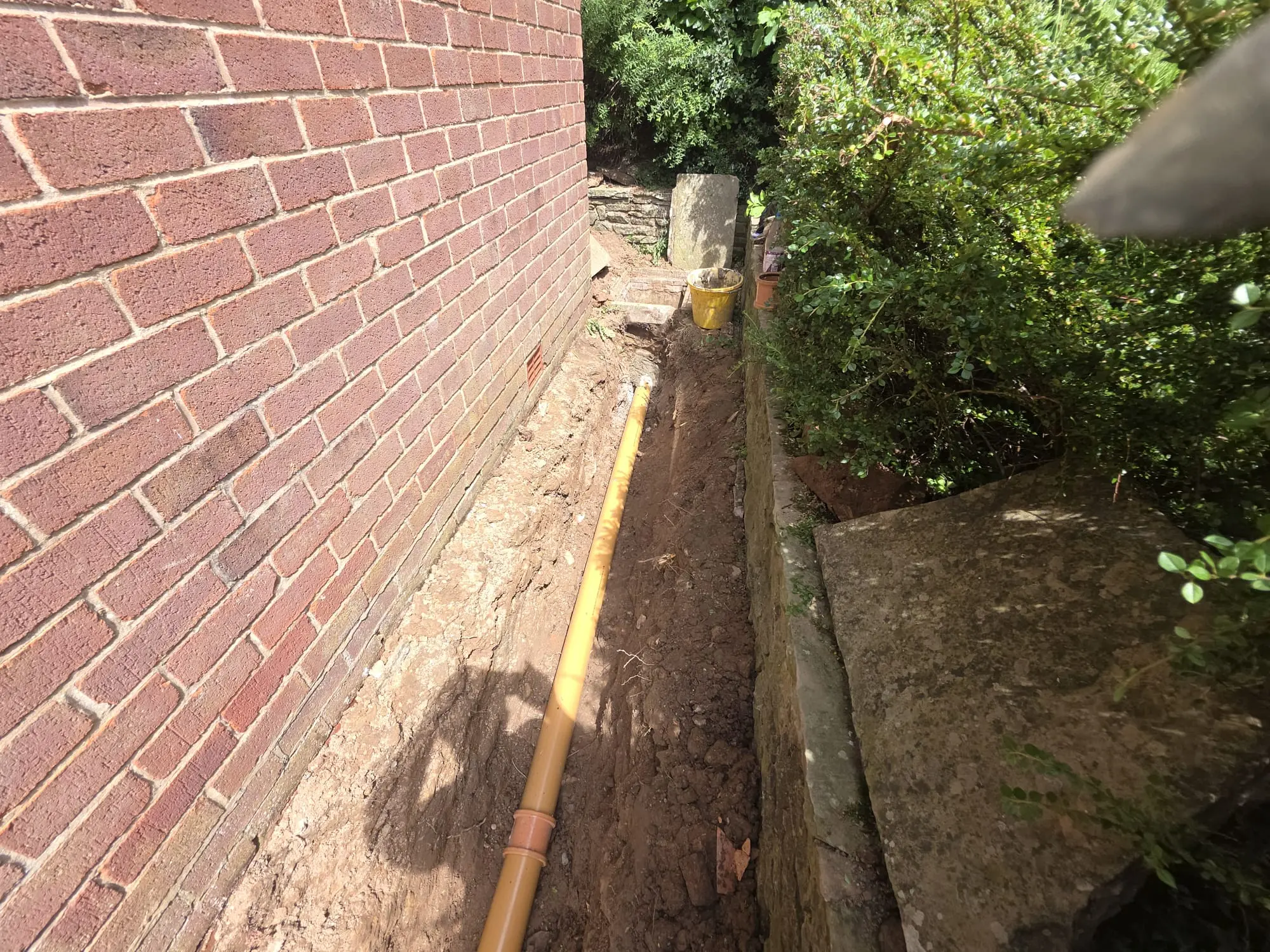 Professional drain excavation and replacement underway, showing a new plastic drainage pipe installed in a trench next to a residential property wall.