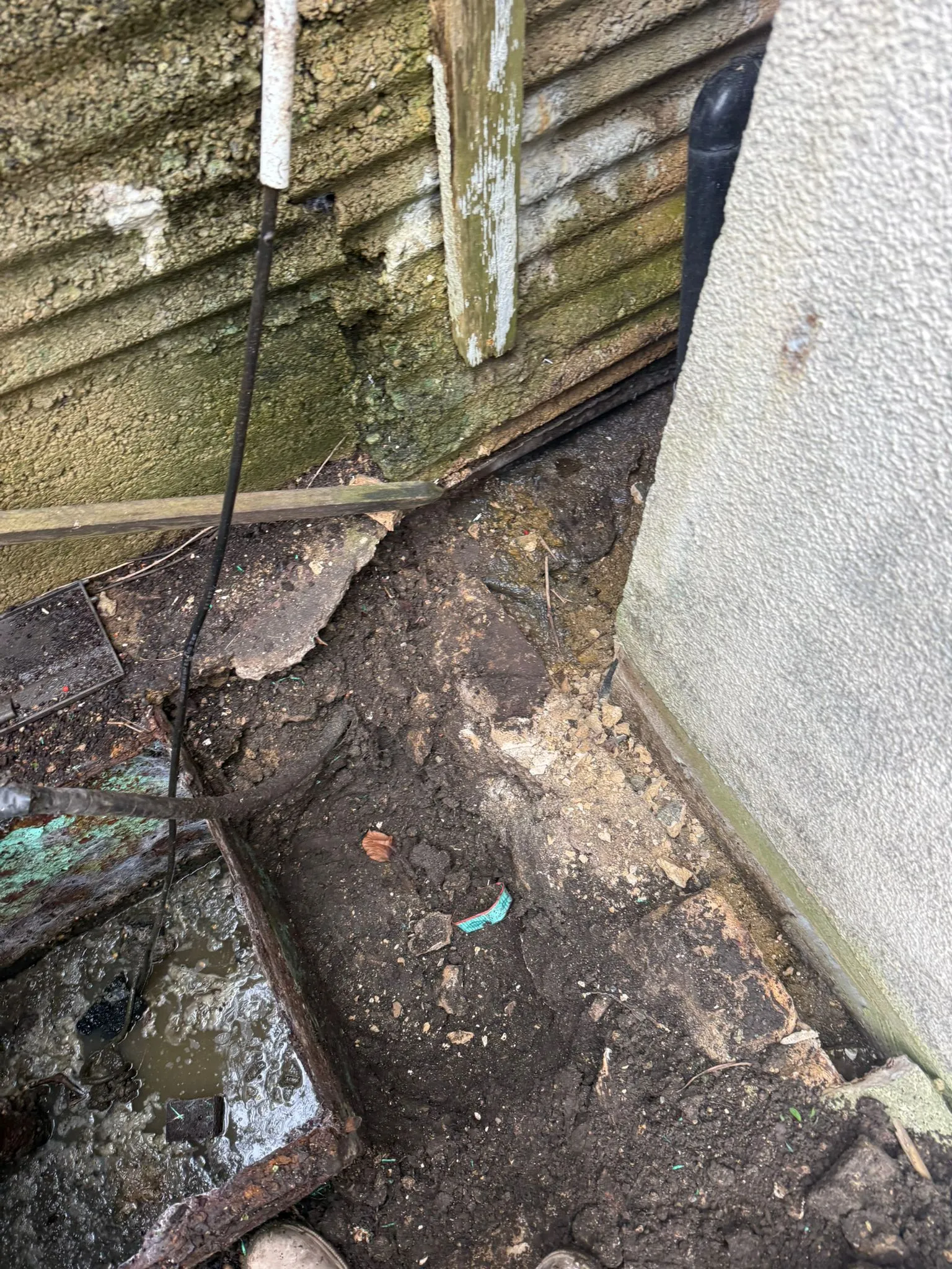 An open, flooded manhole and damaged ground near a building wall, signifying a drainage problem.