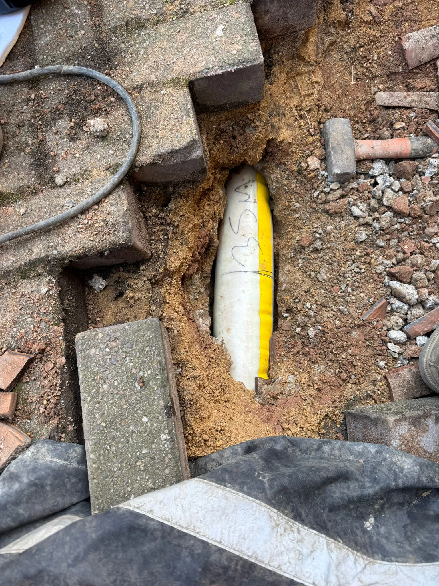 An exposed white and yellow drain pipe revealed through an excavation in a block-paved driveway, showing repair work by Drainage South Ltd.
