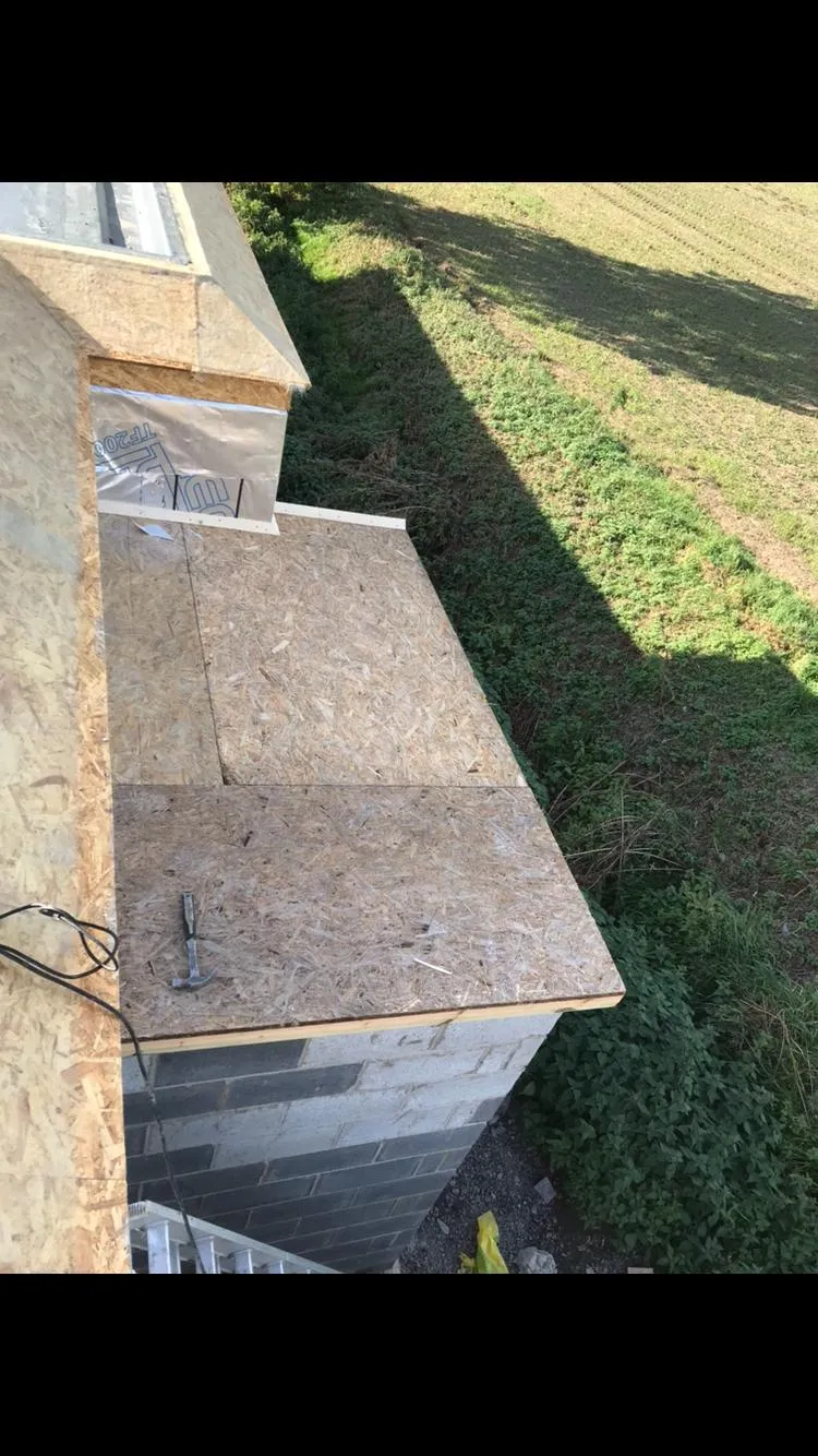 A flat roof deck made of OSB boards under construction, with blockwork walls and a section of pitched roof framing with insulation. A grassy field is visible in the background.