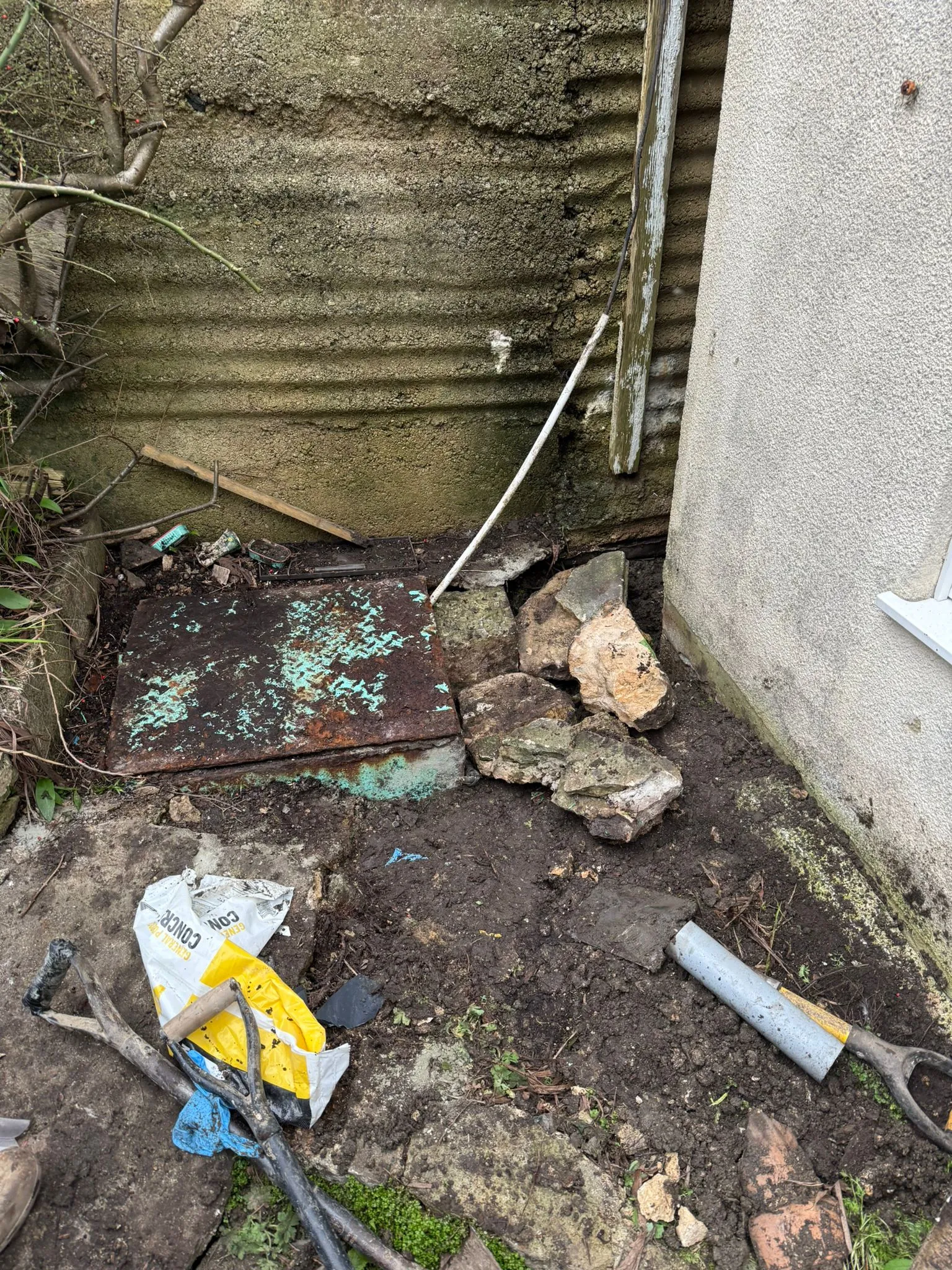 An old metal drain cover next to a house wall surrounded by dug-up earth and shovels, indicating drainage repair work.
