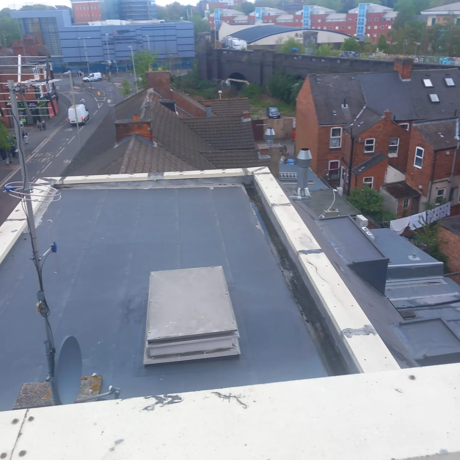 Aerial view of a flat roof with a hatch and antenna, overlooking a busy street with residential tiled roofs, brick houses, and chimneys in urban Leicester.