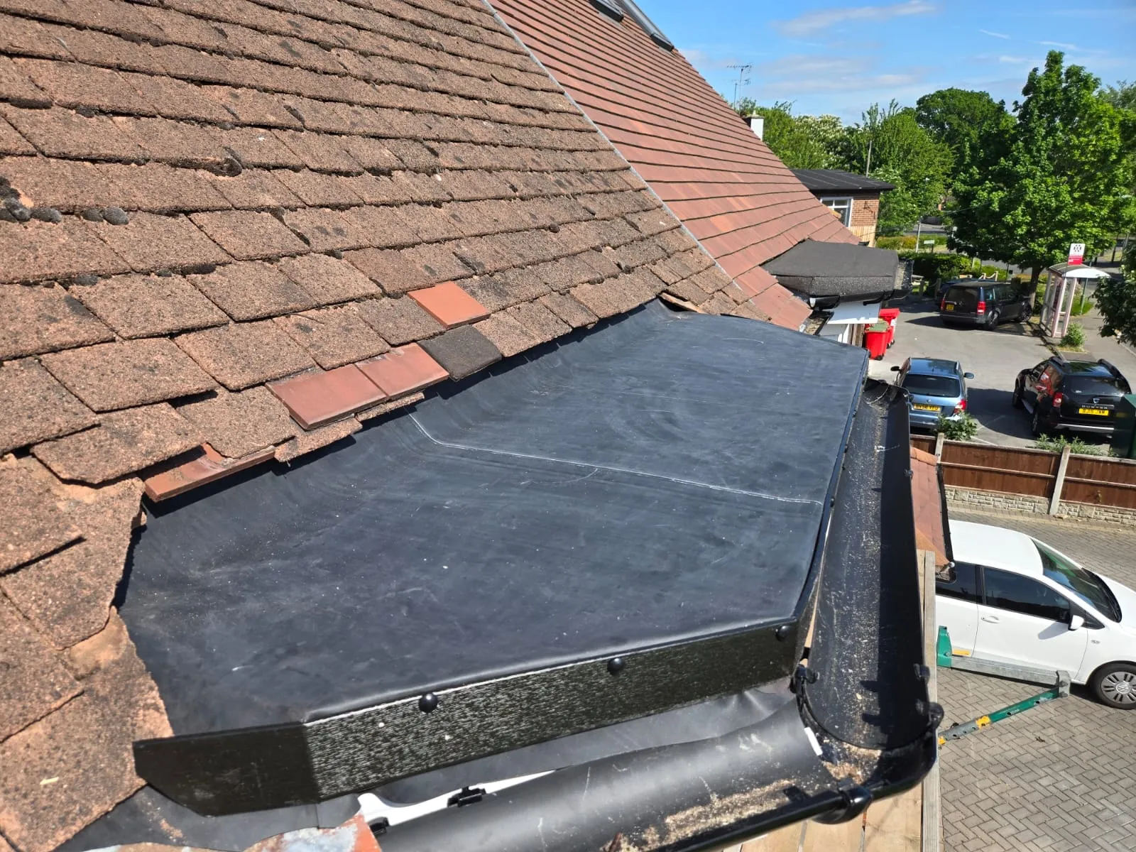 Close-up view of a residential roof showing an older brown tiled pitched roof meeting a dark grey, seamless EPDM flat roof, with black guttering installed.