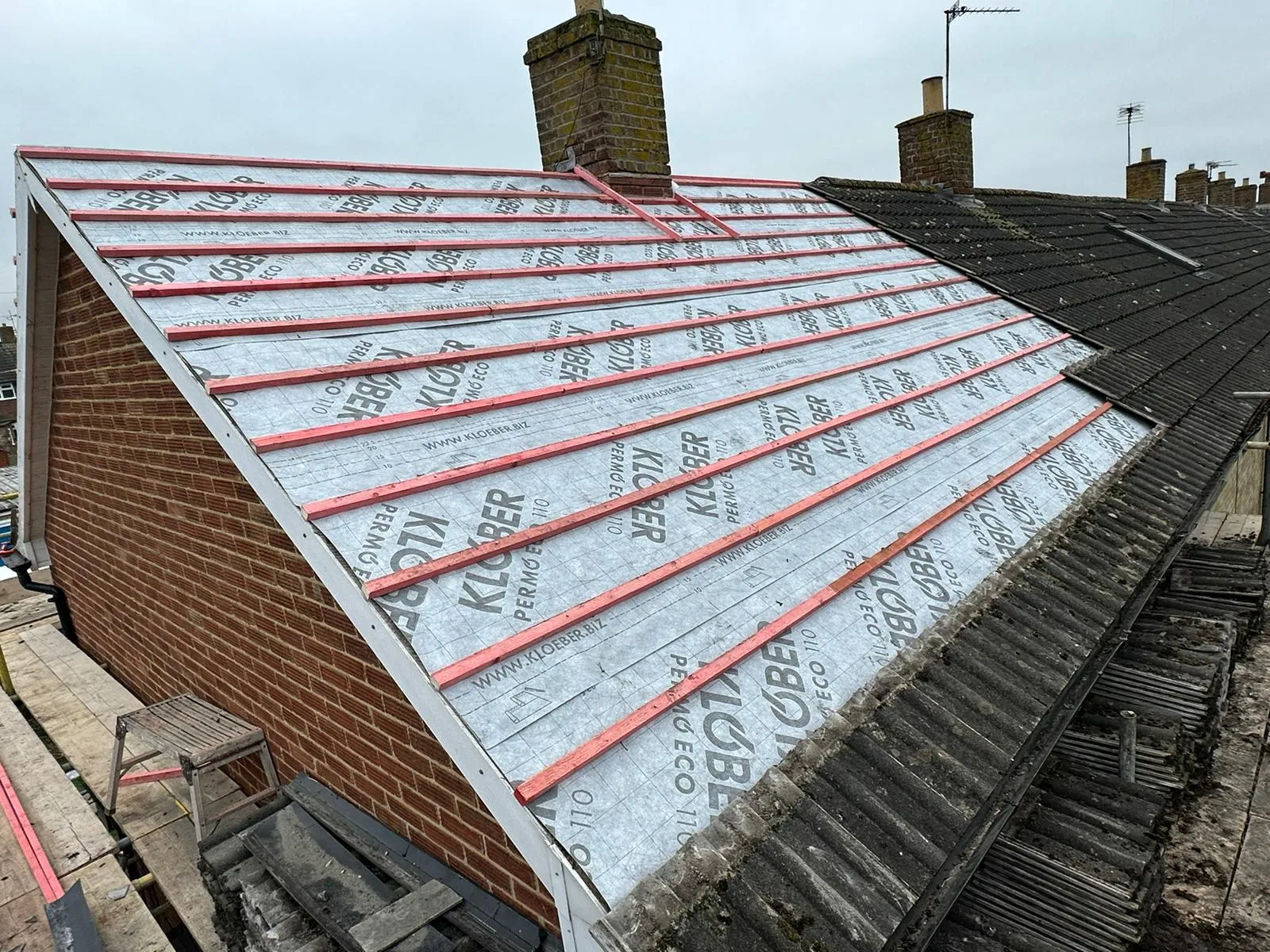A residential roof during replacement, with newly installed underlay and battens on one side and old roof tiles remaining on the other.