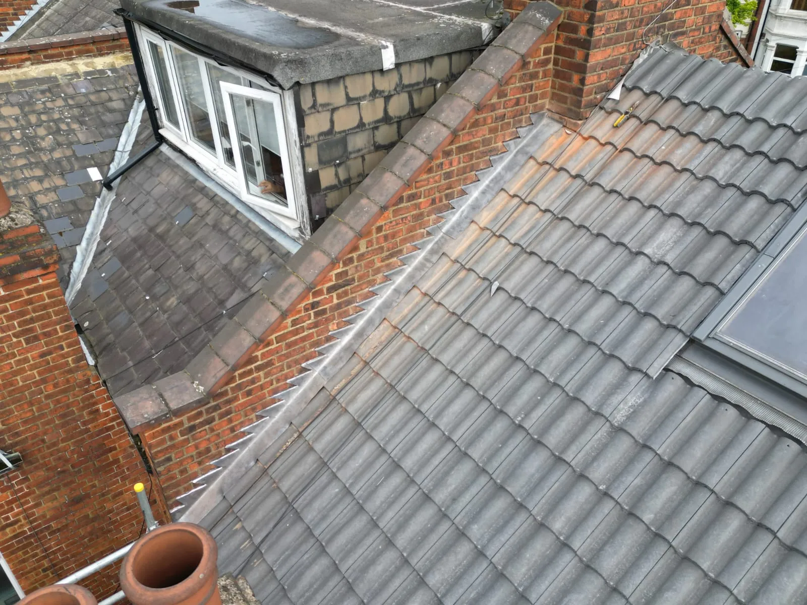 Complex residential roof with grey tiles, a flat roof dormer, brick chimneys, and bird deterrent spikes on lead flashing.
