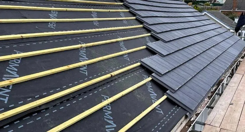 Close-up of a new roof installation in progress, featuring dark grey roof tiles partially installed and roofing underlay with wooden battens ready for further tiling. A complete re-roofing project in Cheshire.