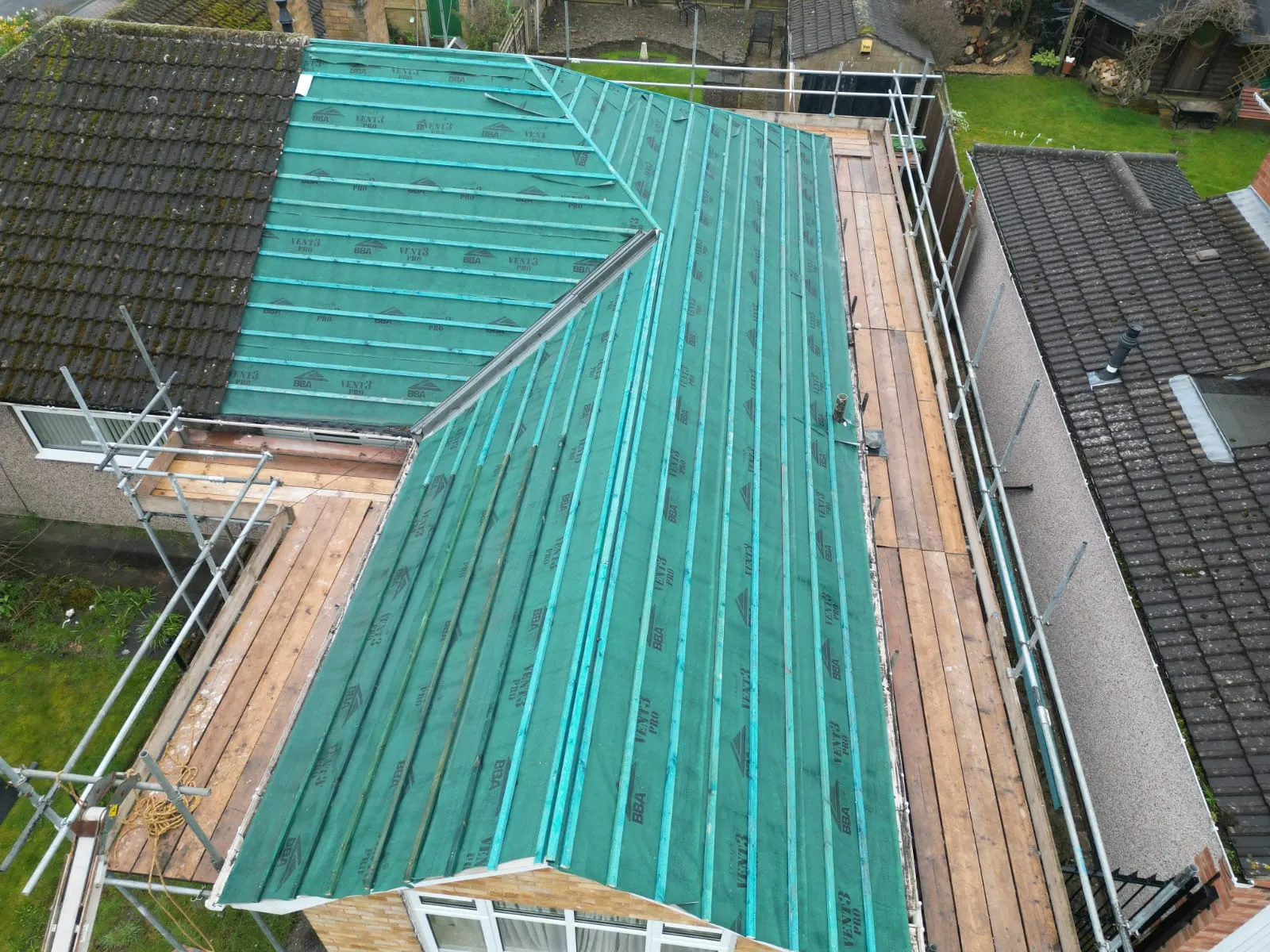 Aerial view of a roof during re-roofing, showing green underlay, wooden battens, and scaffolding.