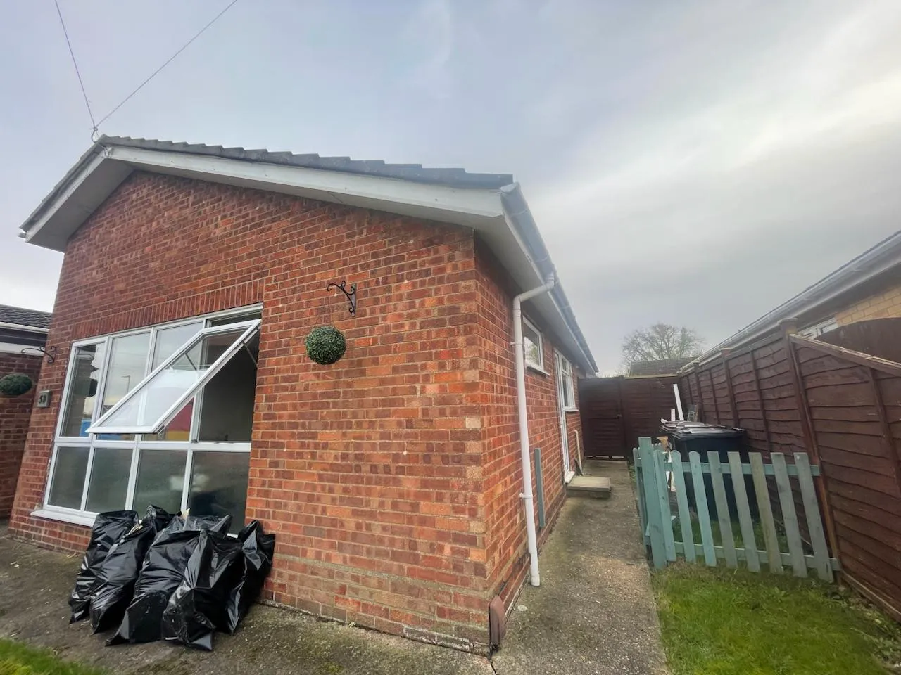 Exterior view of a red brick bungalow with an open window and black waste bags stacked against the wall, indicating domestic asbestos removal or waste disposal.