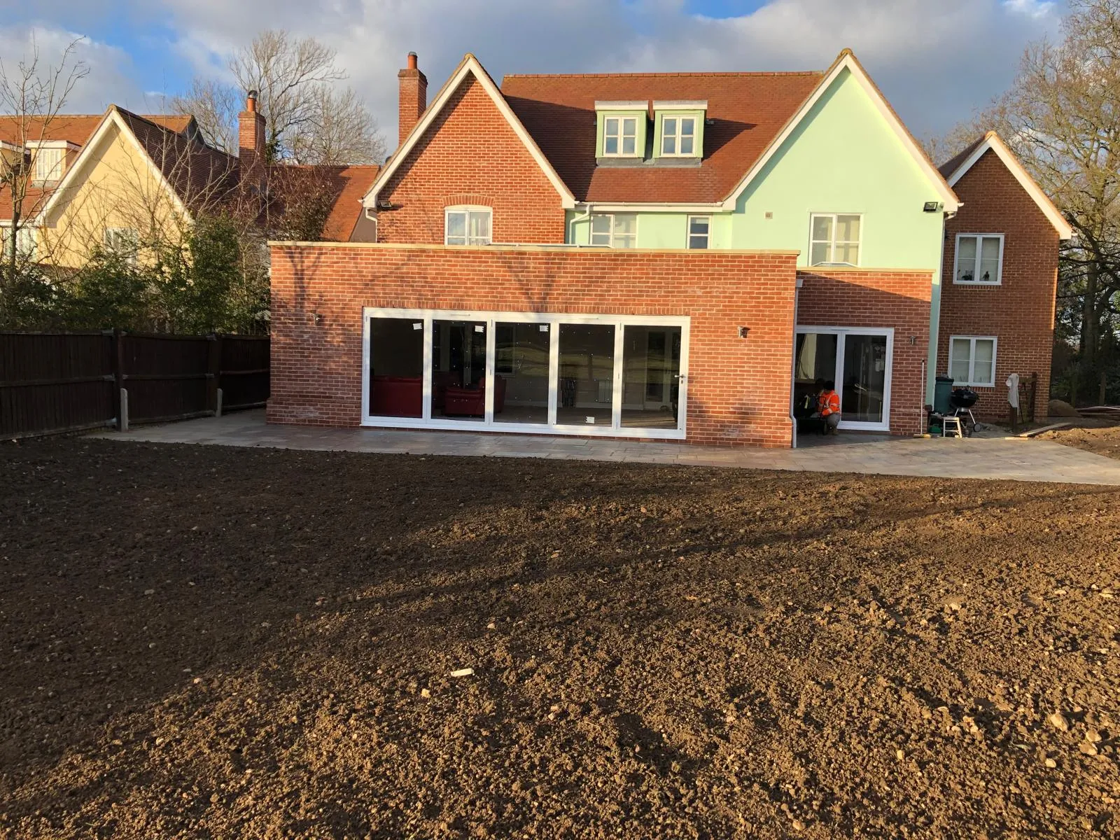 Rear view of a newly built brick house extension with bifold doors and a patio, with an unlandscaped garden in the foreground.