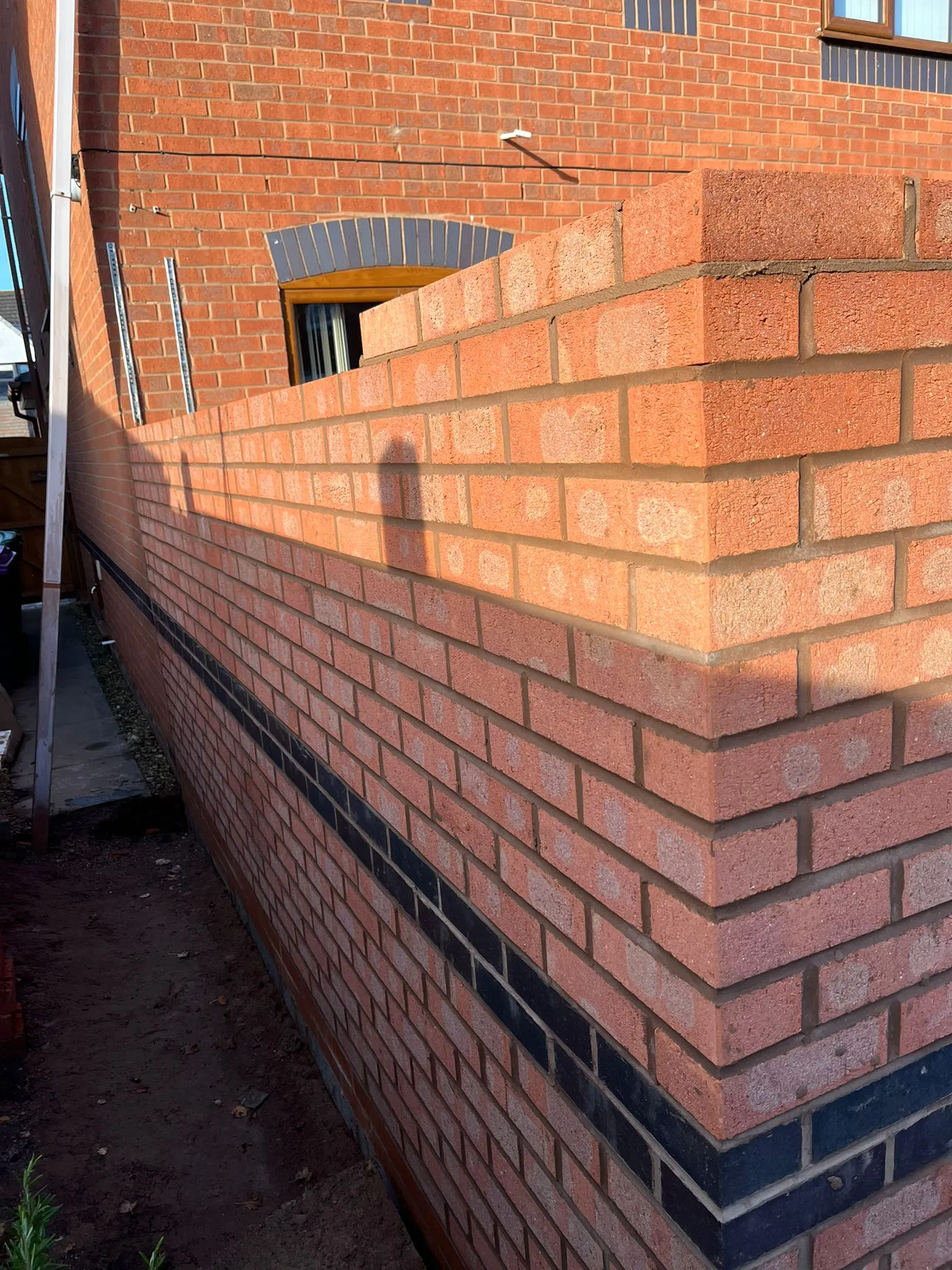A new brick wall under construction, showing the corner of an extension with red and dark blue/black bricks, next to an existing house wall with an arched window.