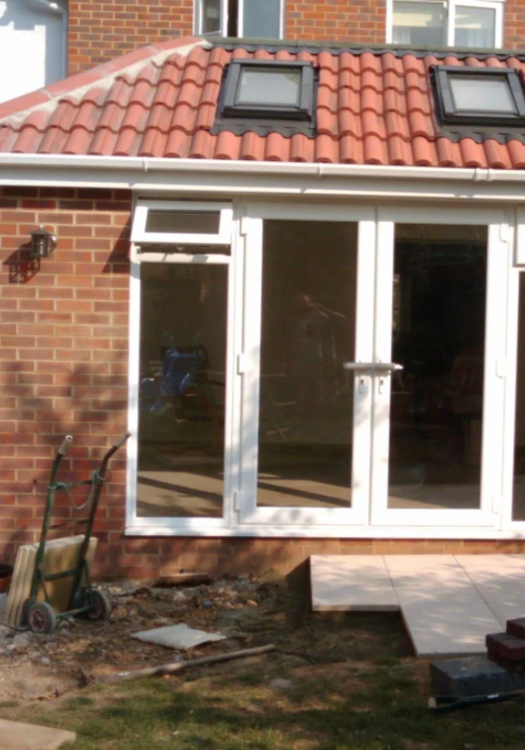 Exterior view of a partial house extension showing a new roof with Velux windows, brick wall, white French doors, and garden landscaping in progress.