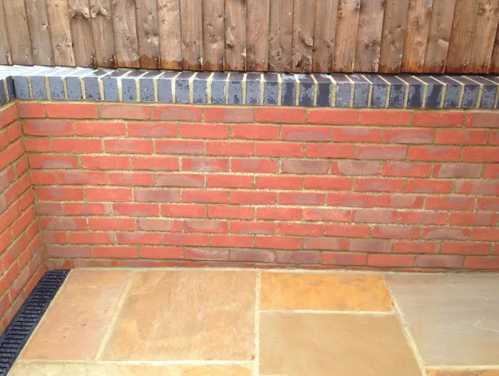 New red brick garden wall with dark coping above a natural stone patio and a linear drain.