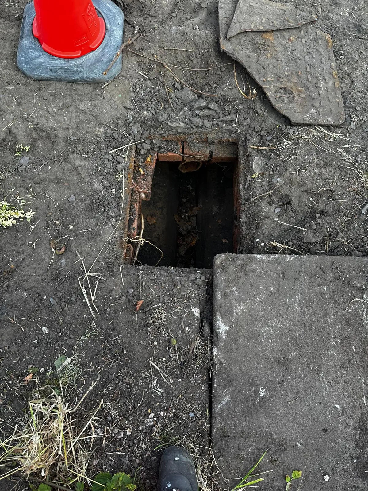An open manhole or inspection chamber in muddy ground with a red cone and concrete slab, revealing brickwork and a drainage pipe containing debris, indicating a problem requiring unblocking or repair.