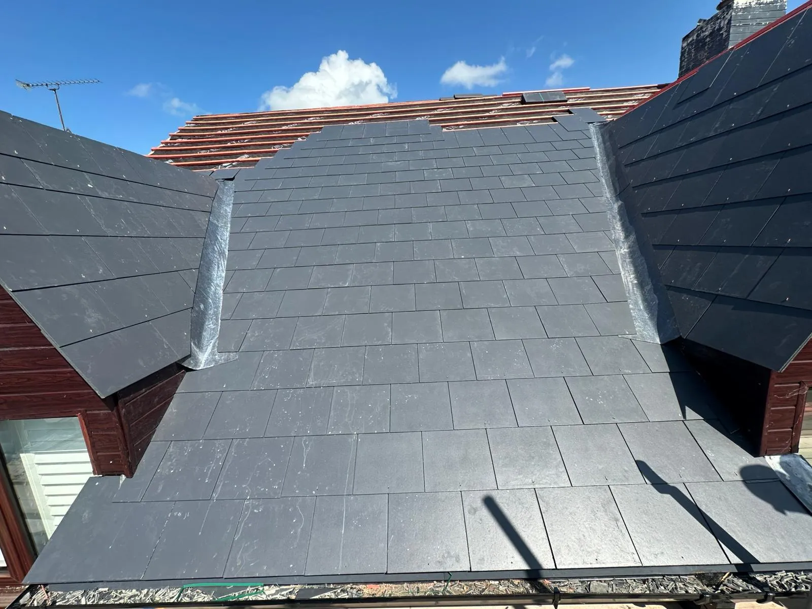 Partial view of a roof being re-tiled with new dark grey slate or tile effect roofing, featuring lead flashing in the valleys and red underlayment visible on the uncompleted section.