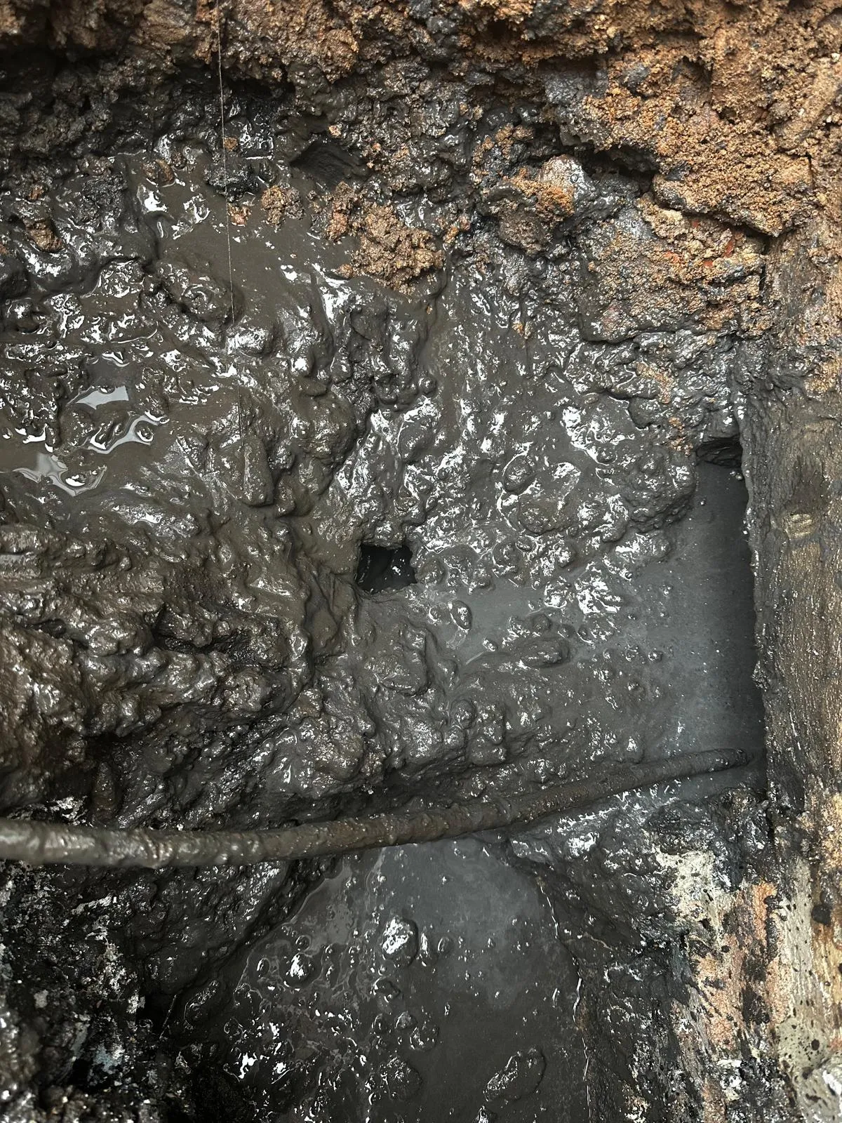 A close-up view of a severely blocked drain, overflowing with dark, greasy sludge and foul water, with a drain rod being used to clear the blockage.