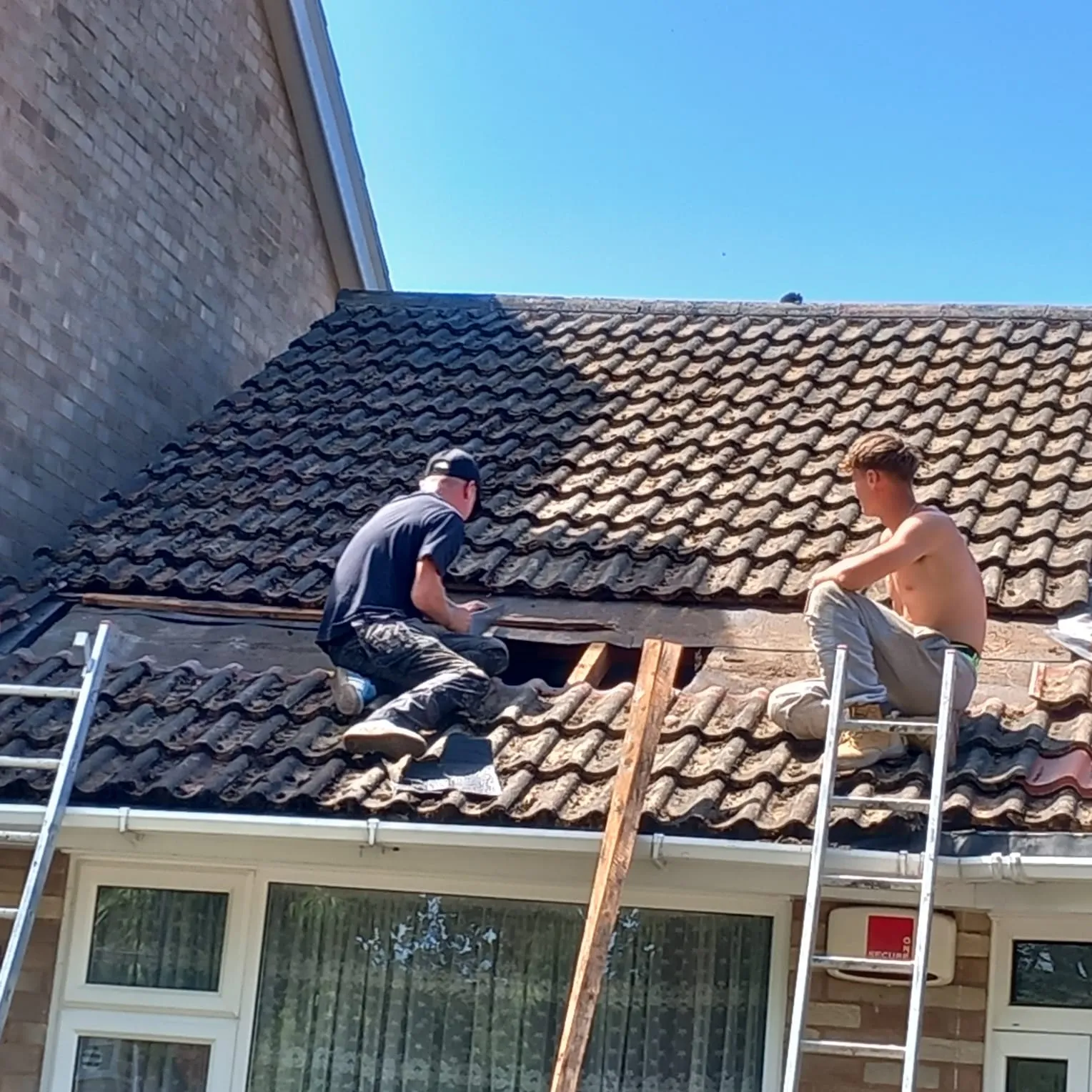 Two skilled roofers actively working on a residential tiled roof, performing a repair or re-roofing service. Parts of the old tiles have been removed.