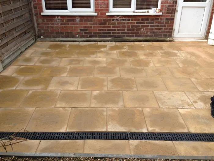 A newly laid light brown patio with drainage channel in front of a brick house wall and fence.