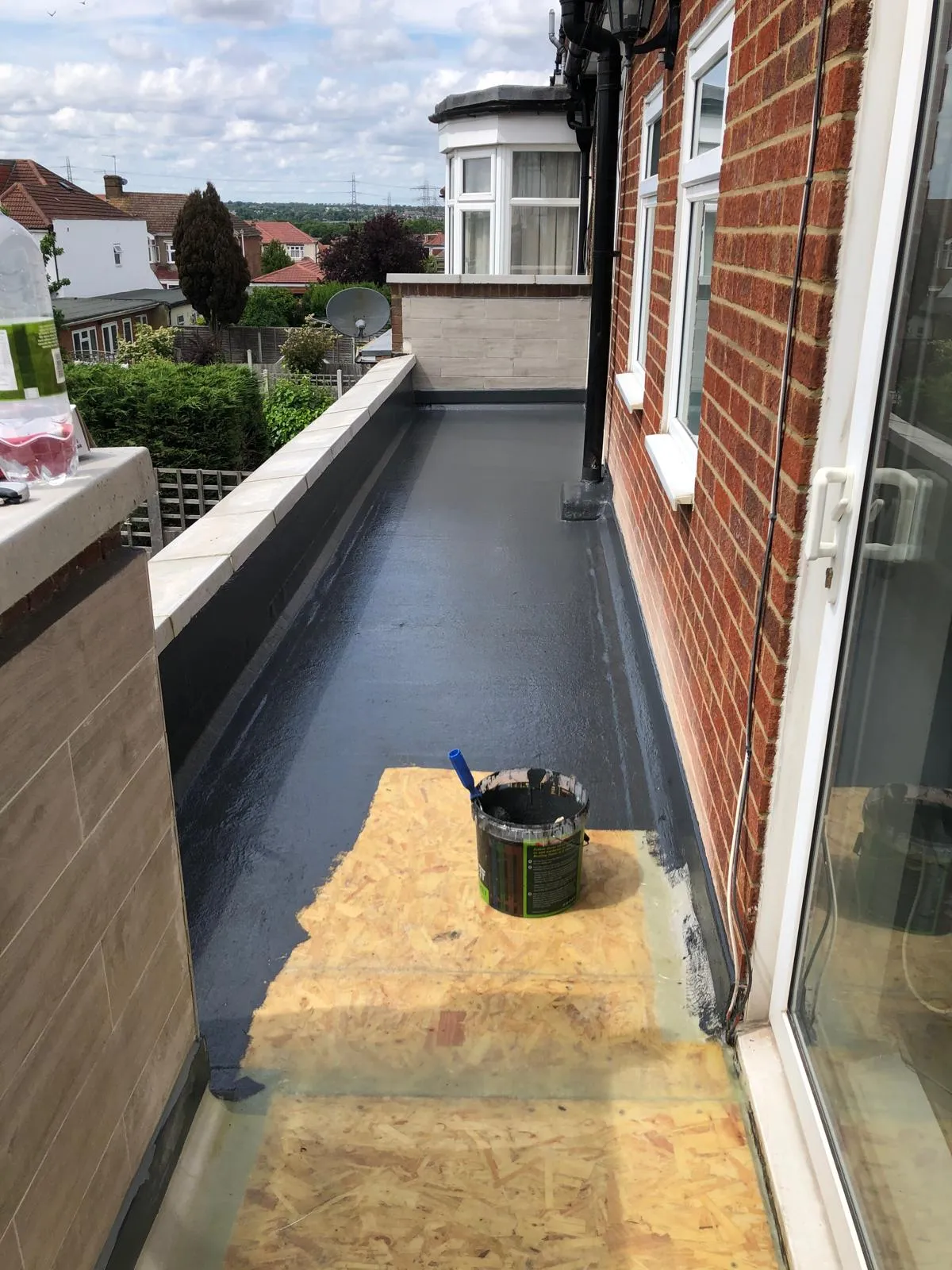 A newly installed or re-coated dark grey flat roof on a residential property, with a bucket of coating and roller visible.