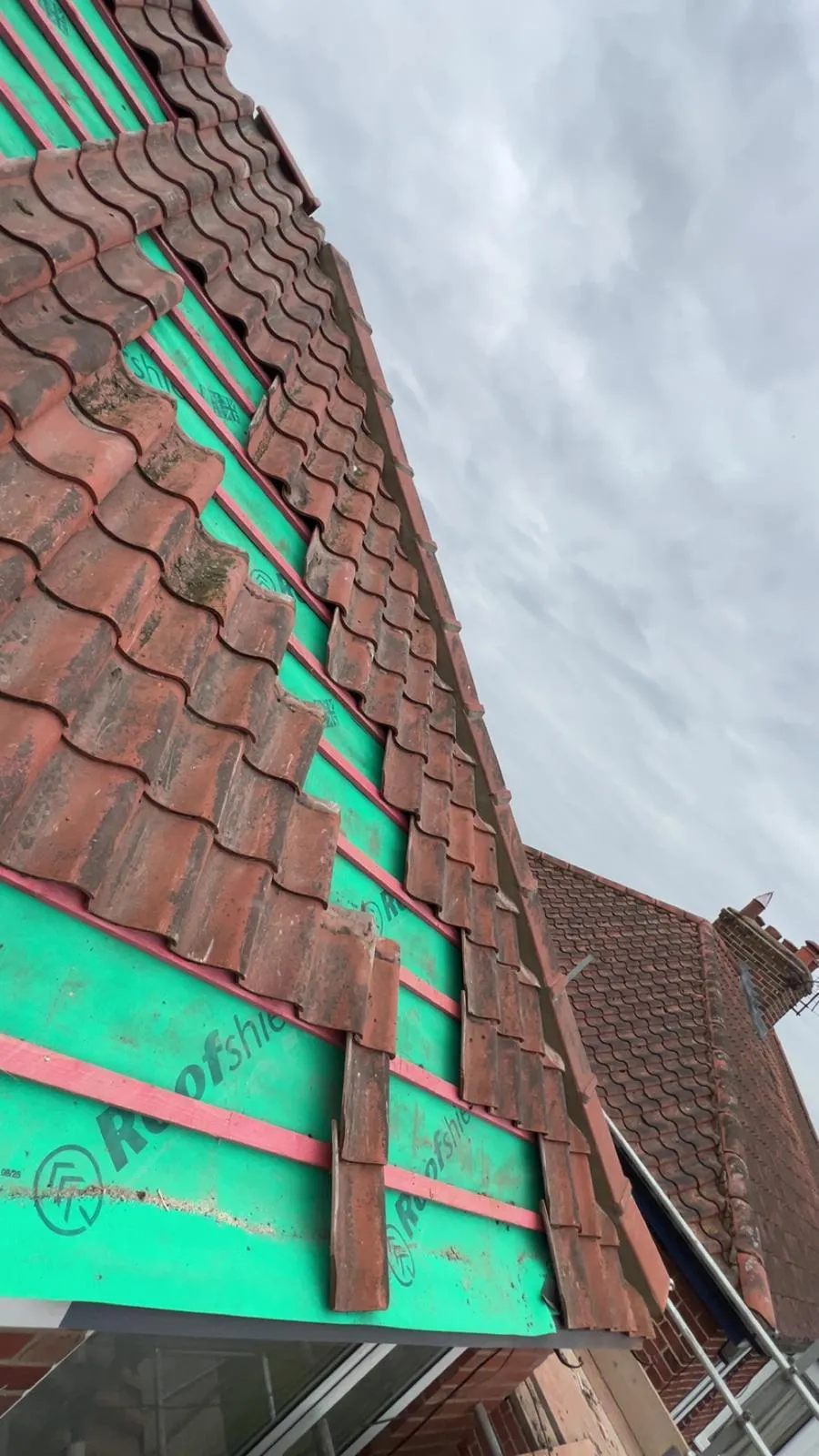 Close-up view of a residential pitched roof with many terracotta tiles removed or in disarray, revealing a green 'Roofshield' underlayment and pink wooden battens during a repair or re-roofing project, set against a cloudy sky.