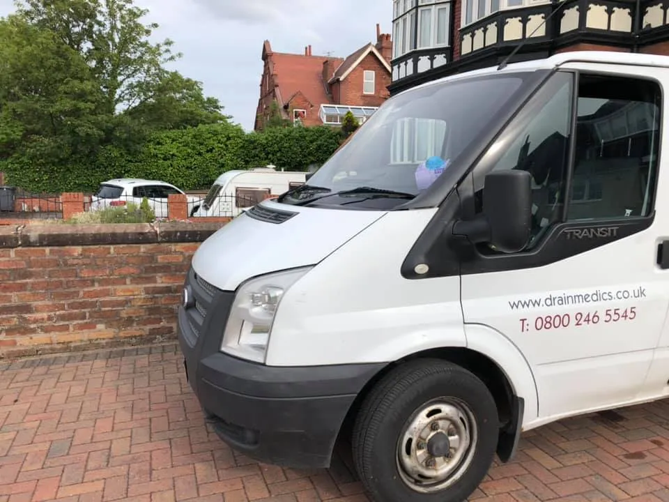 A white drainage service van parked on a paved driveway with a brick wall and residential building in the background.